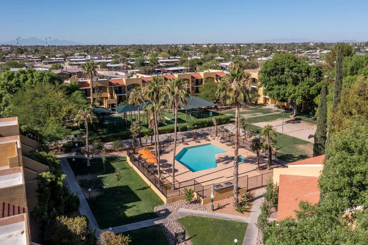 Aerial view of a residential complex featuring a turquoise swimming pool surrounded by palm trees, lounge chairs, and landscaped green areas. In the background, there are several two-story buildings and a tennis court, with a clear blue sky overhead and distant mountains visible.