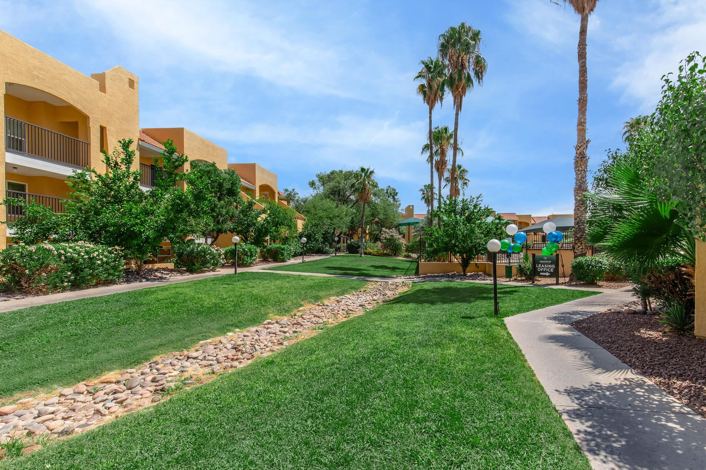 Lush green lawn lined with palm trees, shrubbery, and a rock pathway, leading to two-story yellow buildings. Bright blue sky with scattered clouds enhances the serene atmosphere of the landscaped area. Decorative lights are positioned along paths, inviting a peaceful stroll.