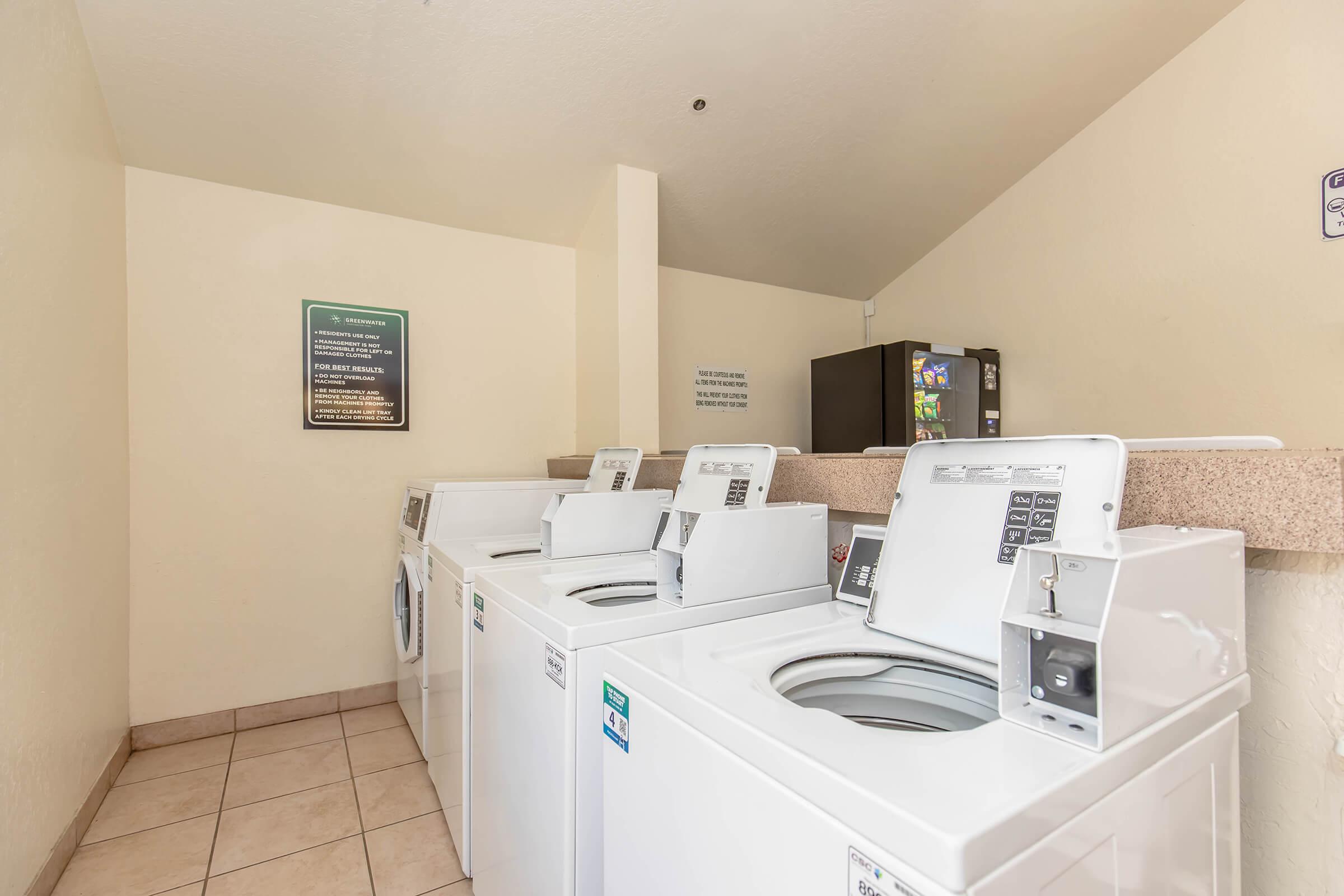 A laundry room featuring several white washing machines lined up against a wall. There is a countertop with a bulletin board above and a refrigerator in the background. The floor is tiled and the walls are painted in a light color, giving the space a clean and organized appearance.