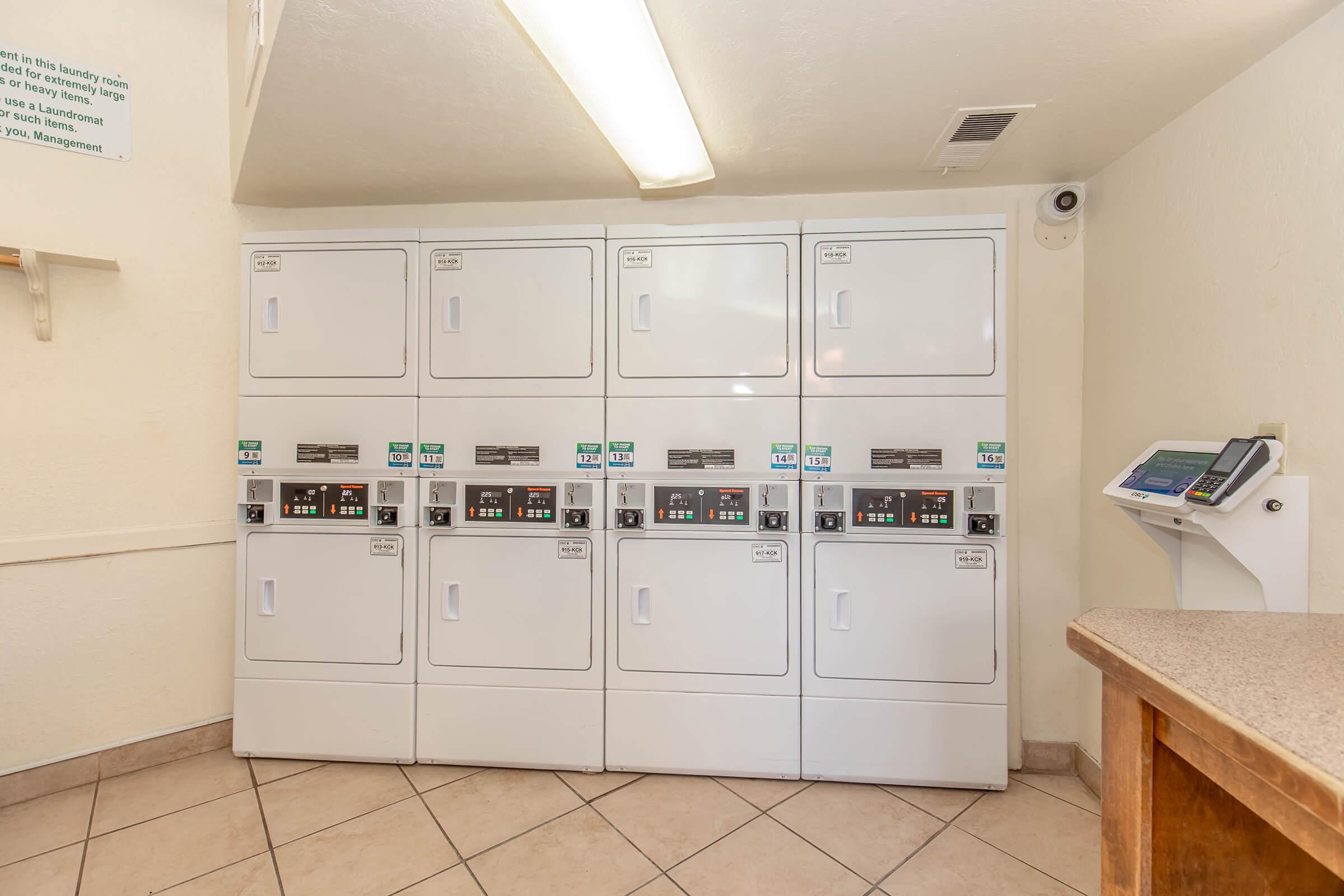 A row of white stacked laundry machines in a laundry room, featuring multiple washers and dryers with digital control panels. A small countertop is visible on the right side, and there’s a payment terminal nearby. The space is well-lit with beige walls and tiled flooring.