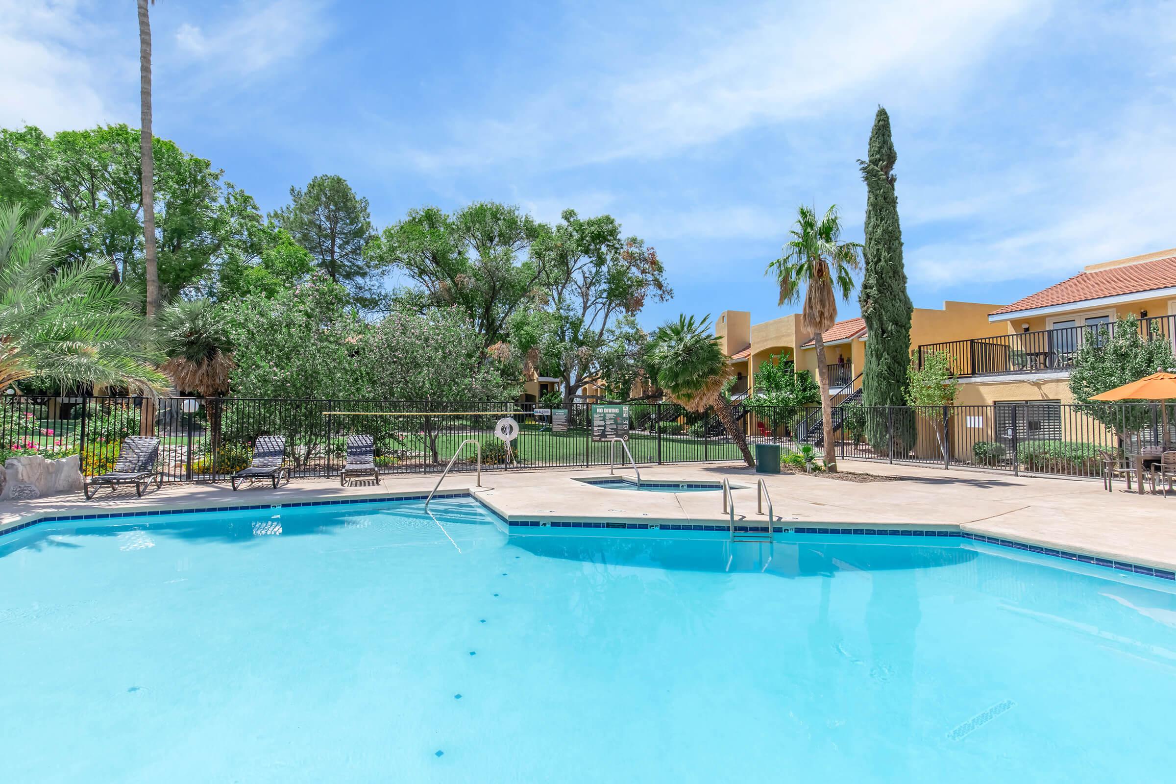 A clear blue swimming pool surrounded by lounge chairs, palm trees, and green landscaping. In the background, there are yellow and orange buildings, adding a tropical ambiance. The sky is bright and sunny, enhancing the relaxing atmosphere of the outdoor space.