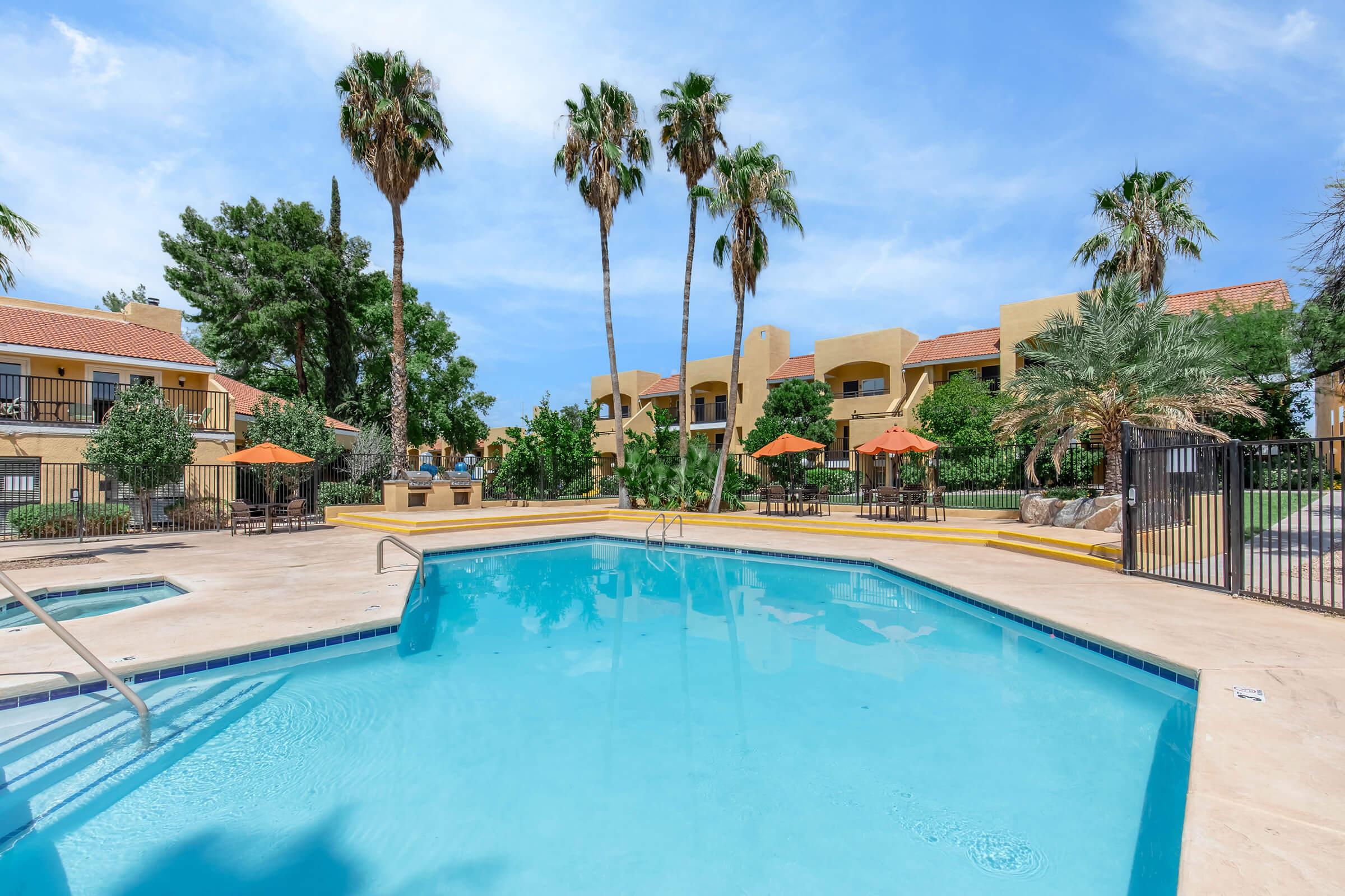 A clear blue swimming pool surrounded by palm trees and lush greenery. In the background, there are yellow buildings with balconies and orange umbrellas set up for shade. The scene is bright and inviting, showcasing a relaxing outdoor space ideal for leisure and enjoyment.