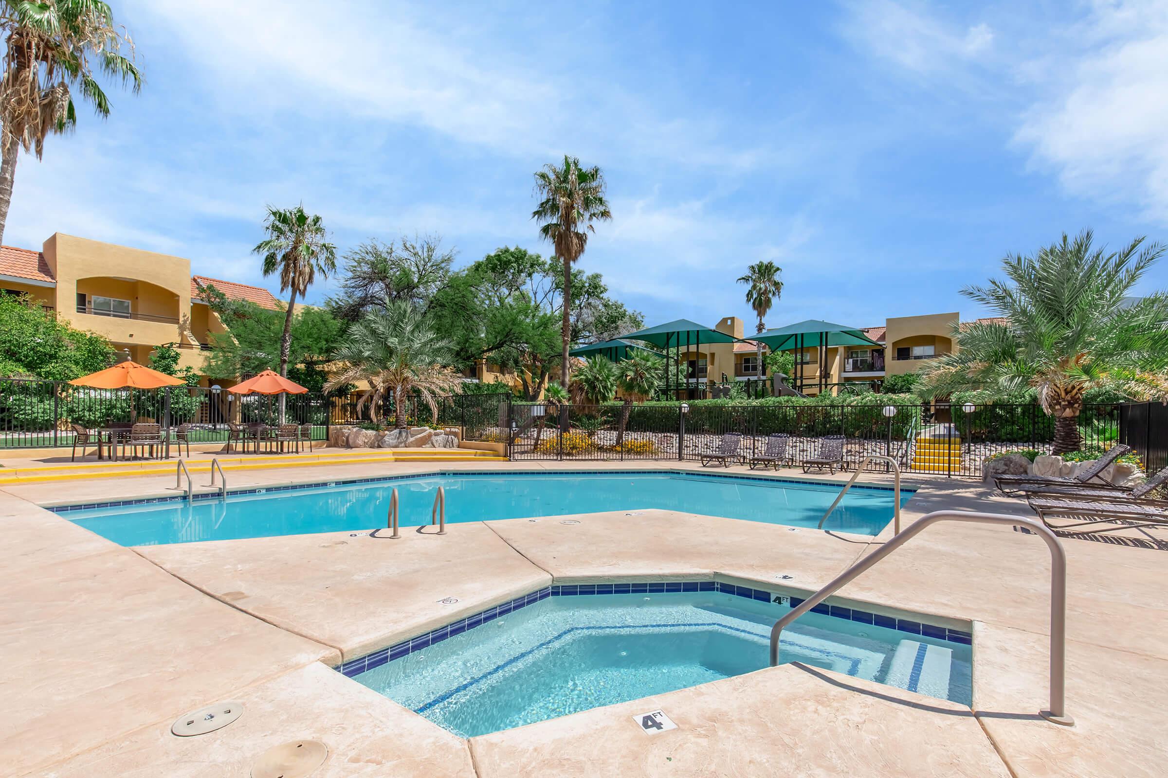 A sunny pool area featuring a rectangular swimming pool and a hot tub. Surrounding the pool are lounge chairs and orange umbrellas, with palm trees and lush greenery in the background. Residential buildings are visible in the distance under a clear blue sky.