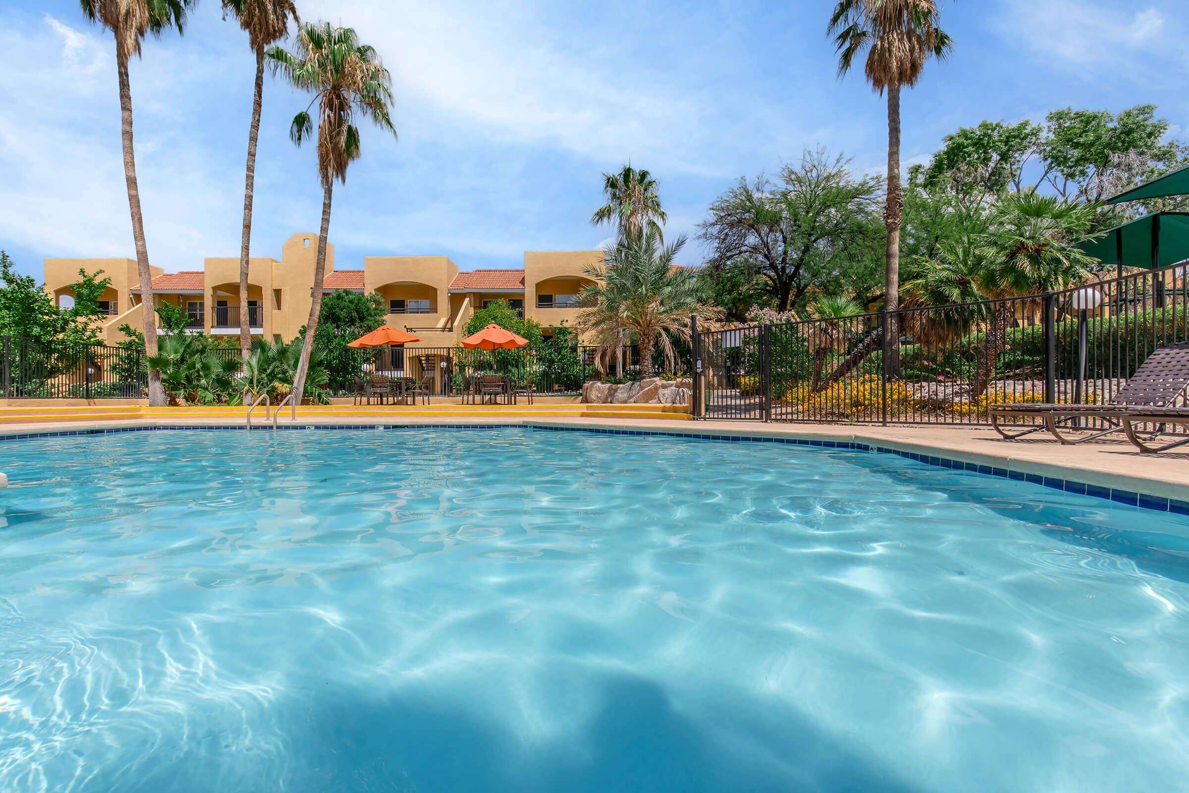A clear blue swimming pool with rippling water in the foreground. In the background, lush green palm trees and colorful buildings are visible, along with orange umbrellas and lounging chairs around the pool area. The sky is bright and partly cloudy, creating a vibrant, inviting atmosphere.