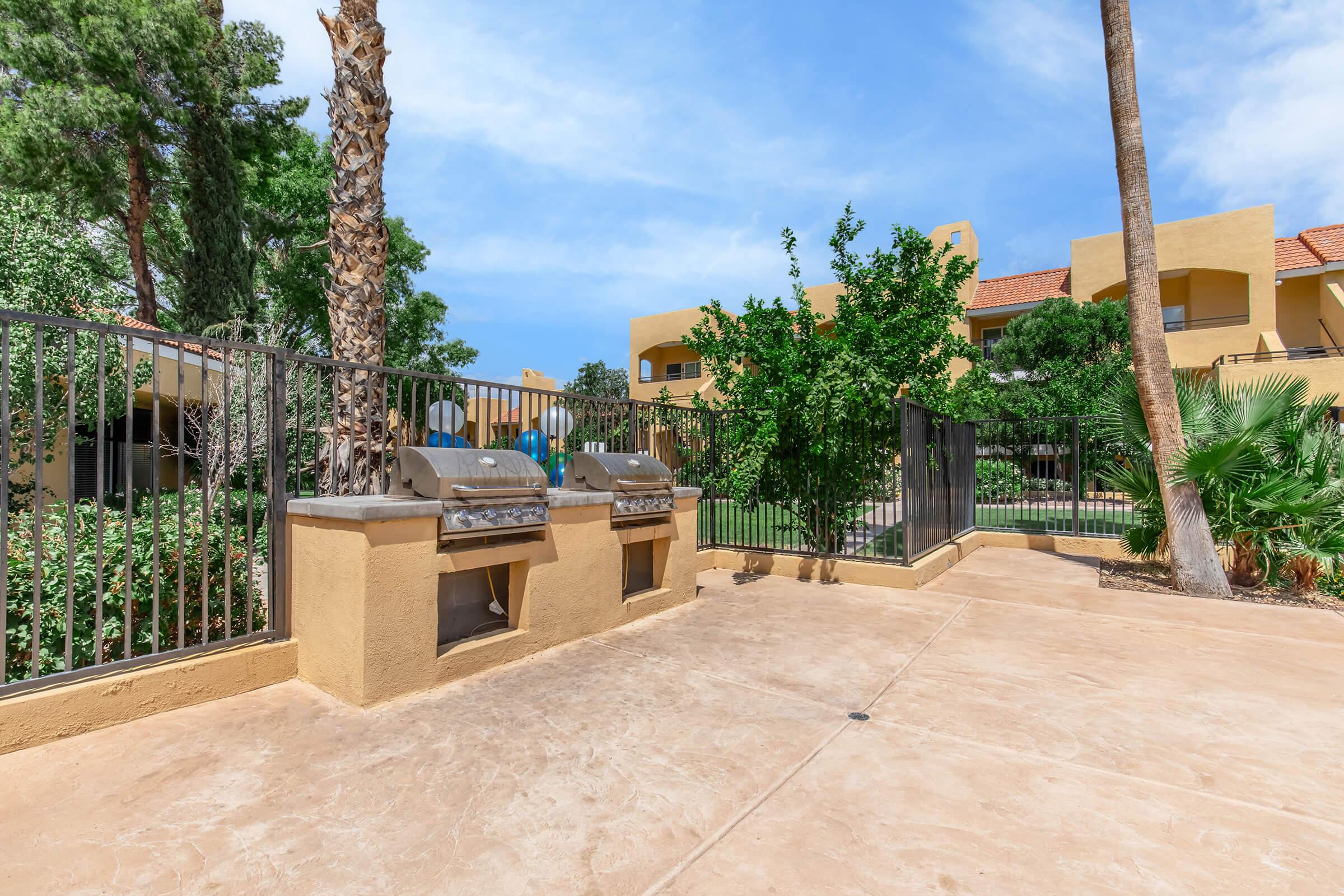 Two outdoor grills on a concrete patio surrounded by palm trees and greenery. In the background, there are residential buildings with yellow walls and red tile roofs, under a blue sky with scattered clouds. The area is enclosed by a black fence, creating a private outdoor space.