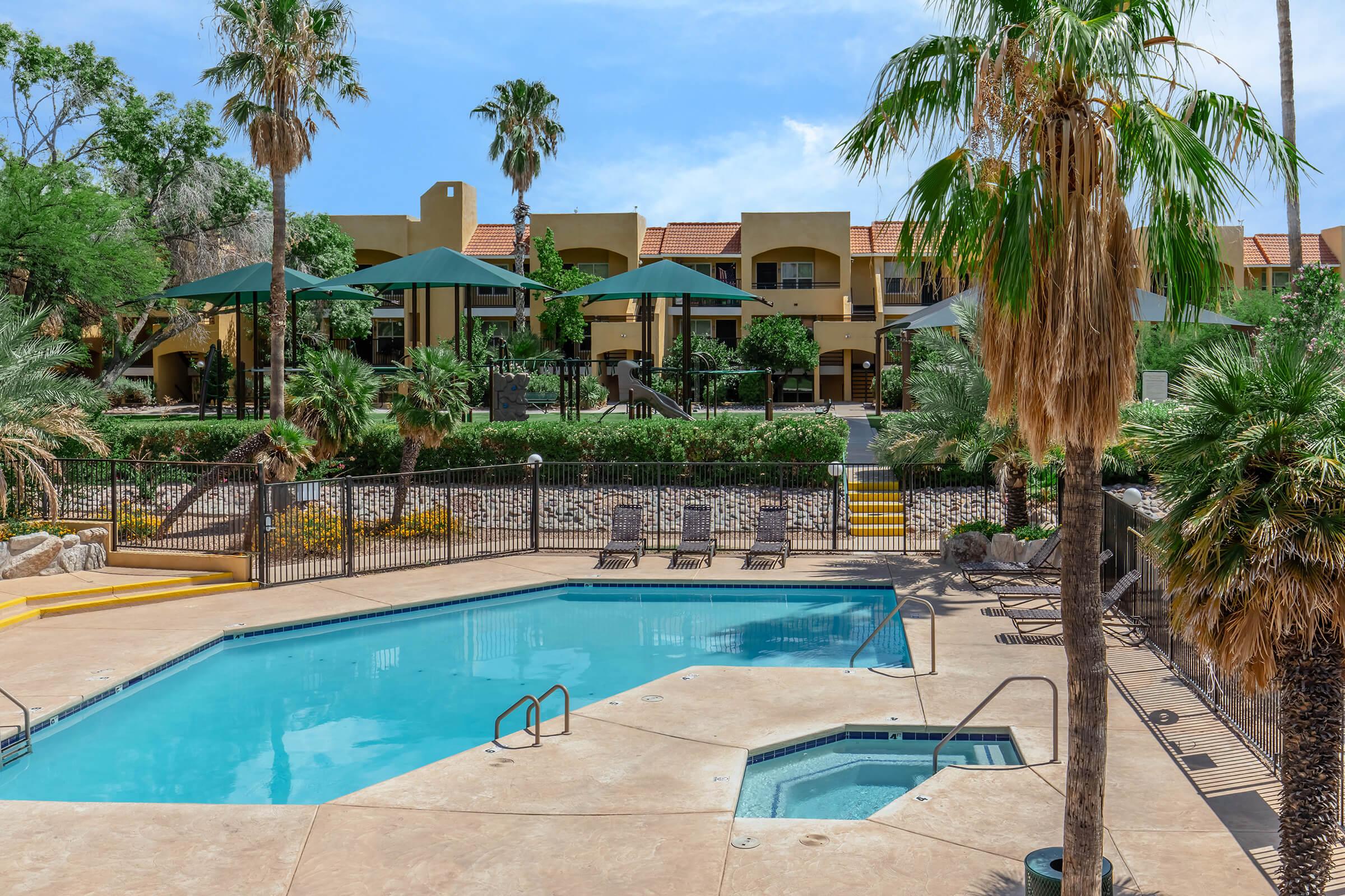 A serene outdoor pool area surrounded by palm trees and lounge chairs. The pool features a shallow entry and a hot tub nearby. In the background, there are two-story buildings with balconies, shaded by green umbrellas. The scene captures a relaxed atmosphere in a sunny environment.
