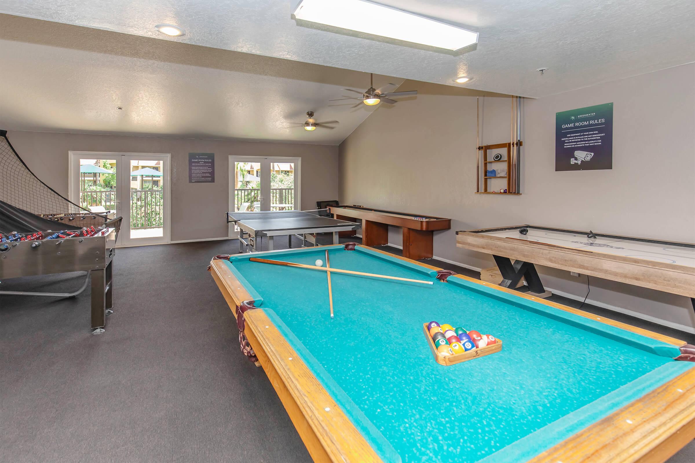 A game room featuring a green pool table with a cue stick and balls, a foosball table, a ping pong table, and large windows allowing natural light. The decor is modern, with a neutral color scheme and ceiling fans. There are activity rules displayed on the wall.
