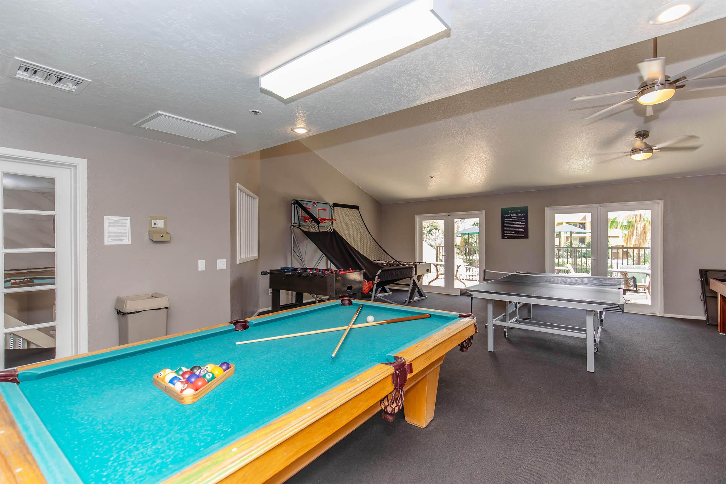 A game room featuring a billiard table with a triangle of pool balls, a foosball table, a basketball arcade game in the background, and a ping pong table. The room has neutral-colored walls, a ceiling fan, and ample natural light from windows.