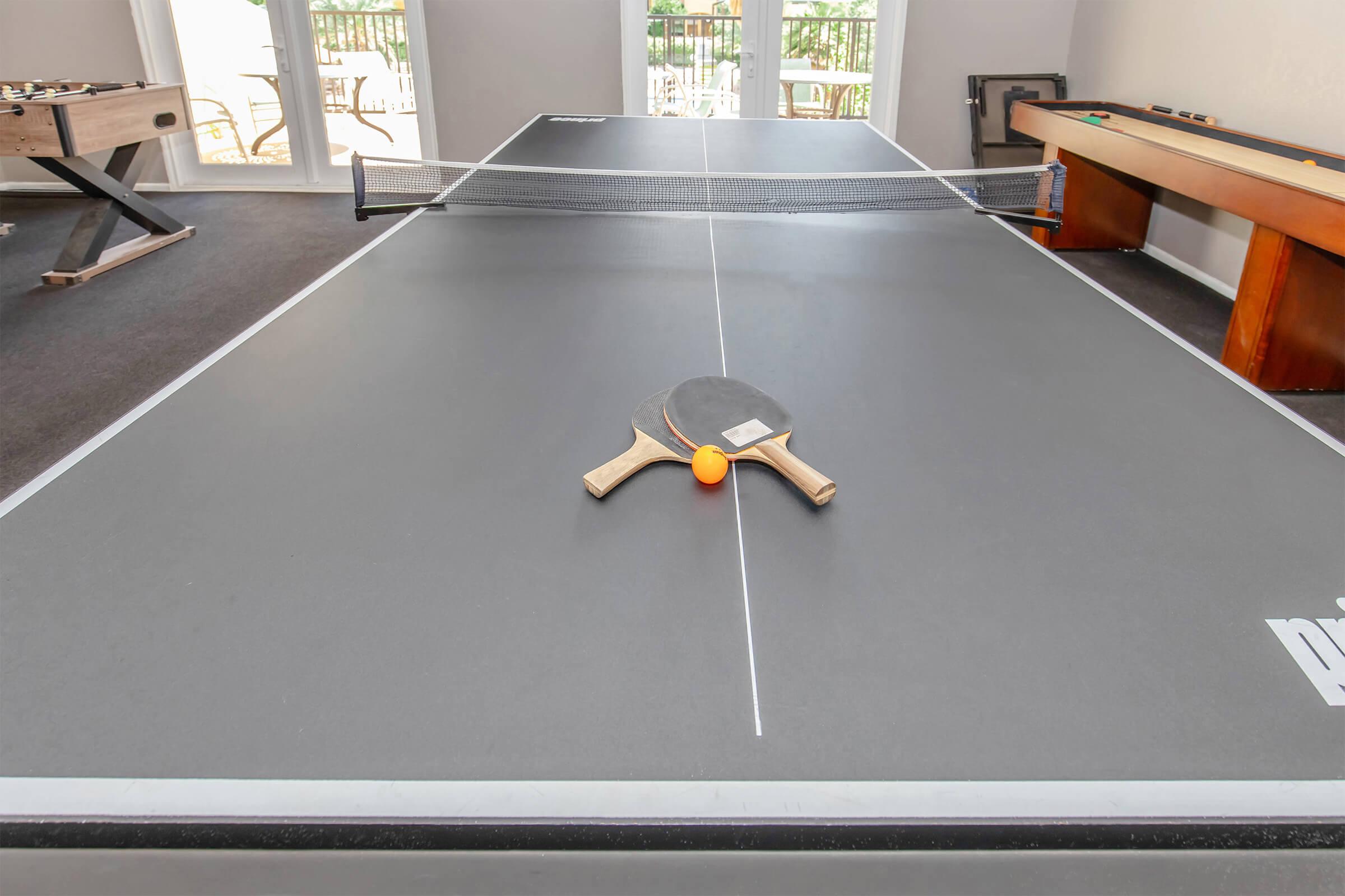 A ping pong table featuring a black surface, with a paddle and an orange ball positioned at the center of the table. The backdrop includes windows allowing natural light, enhancing the lively atmosphere of the recreational space.