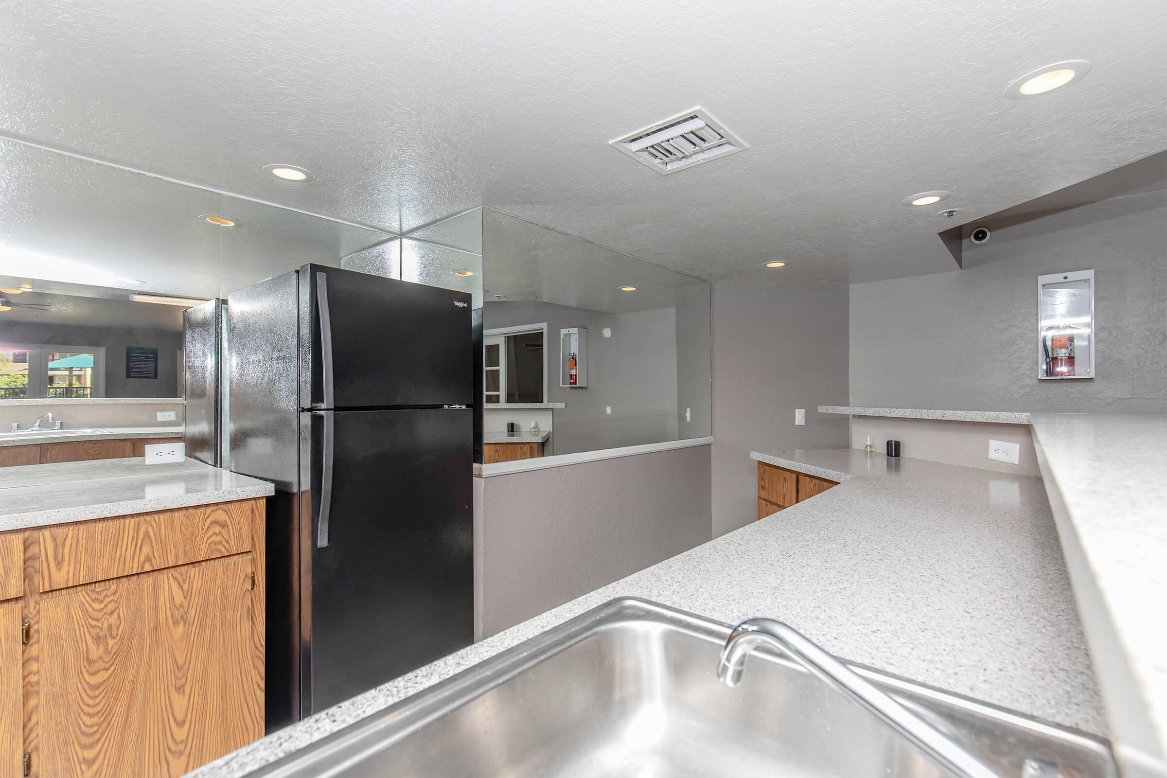 A modern kitchen featuring dark wooden cabinets, a stainless steel sink, and a black refrigerator. The walls are a light gray, and there are multiple light fixtures overhead. A large mirror reflects the countertop and adds depth to the space, creating a bright and airy feel.