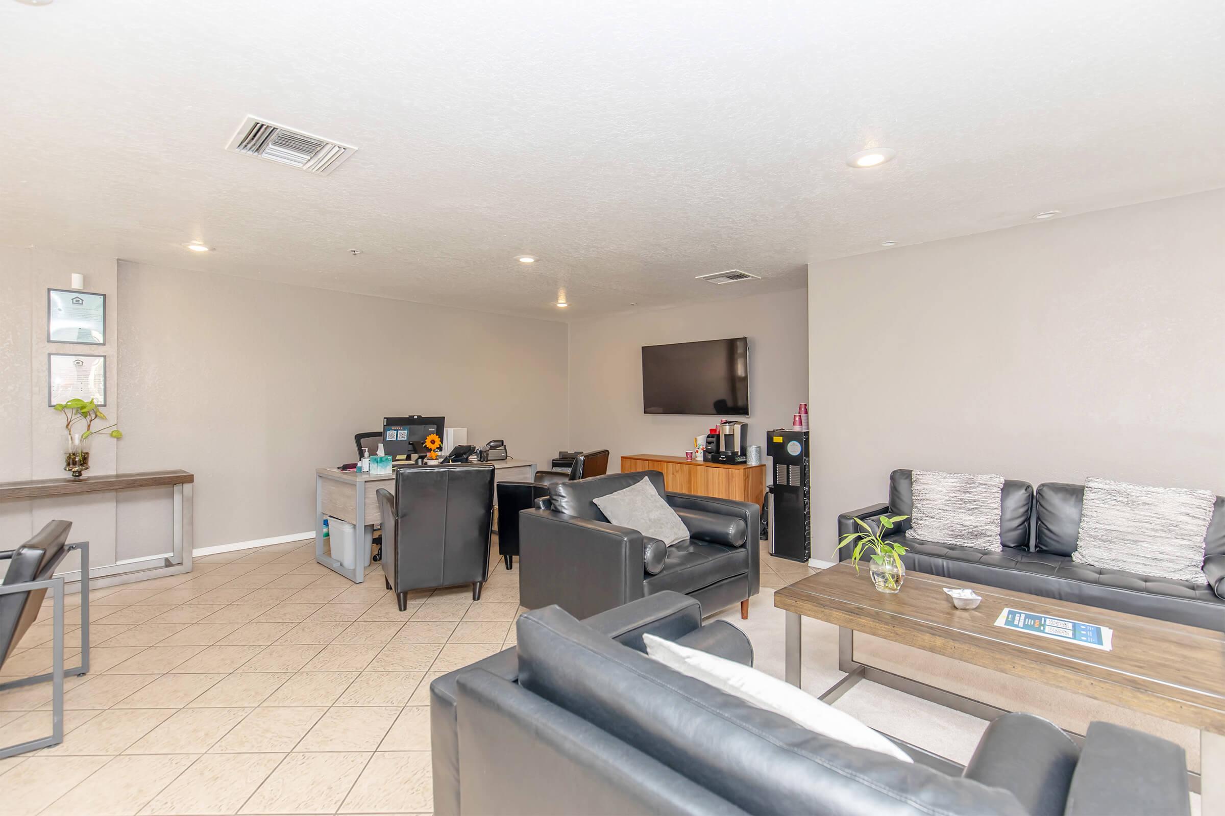 A modern, well-lit lounge area featuring black leather sofas, a wooden coffee table, and a wall-mounted television. A desk with office supplies is visible in the background, along with decorative plants. The space has beige walls and tiled flooring, creating a welcoming atmosphere.