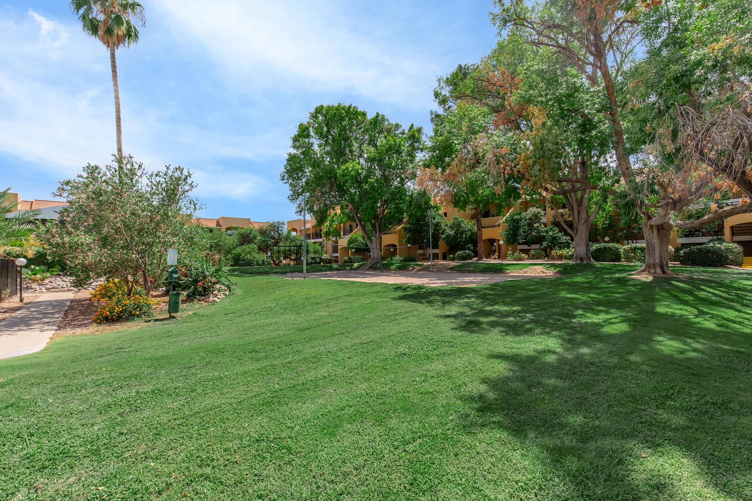 Lush green grass in a landscaped area with trees and shrubs, featuring a pathway and residential buildings in the background. Bright blue sky with a few clouds and a palm tree visible. The scene conveys a tranquil outdoor environment.