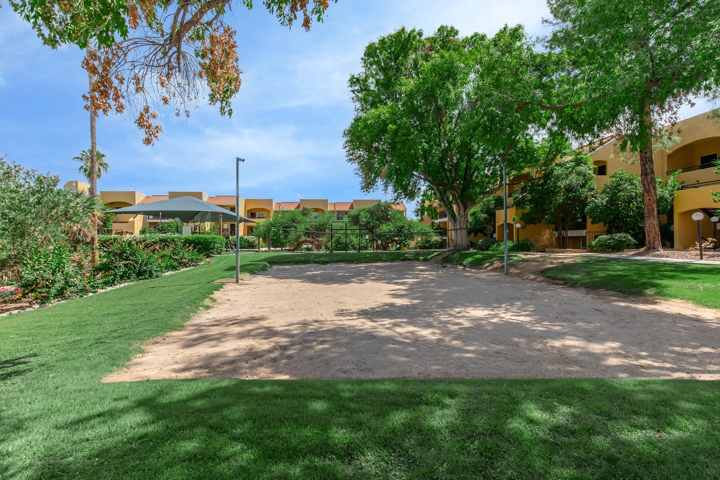 A sunny outdoor area featuring a sandy volleyball court surrounded by lush greenery, trees, and landscaped gardens. Residential buildings are visible in the background, providing a serene and inviting atmosphere.