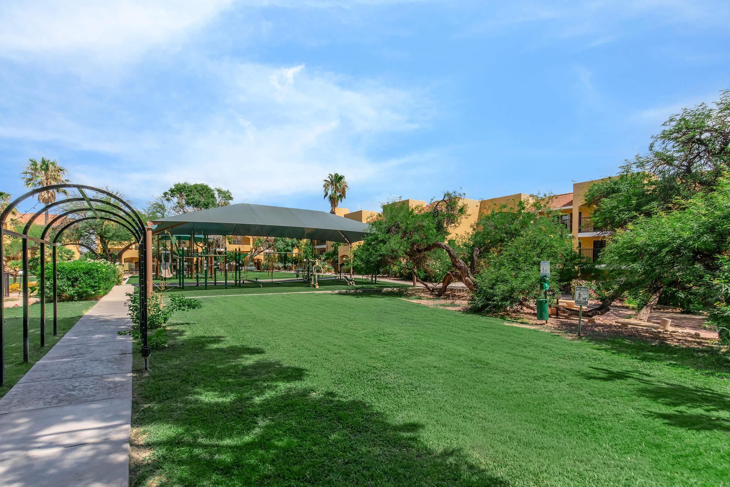A serene green park area featuring lush grass and shaded play structures. Surrounding trees provide a natural ambiance, while nearby buildings add a touch of color to the scene. The sky is clear and blue, creating a bright and inviting atmosphere for outdoor activities.