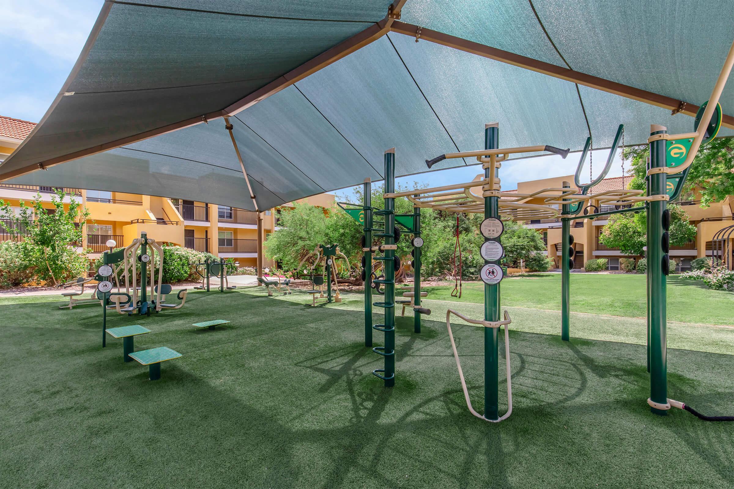 Outdoor fitness area covered by a shade structure, featuring various exercise equipment such as pull-up bars, climbing ladders, and benches. The space is surrounded by greenery and is part of a residential complex, providing a recreational environment for physical activities.