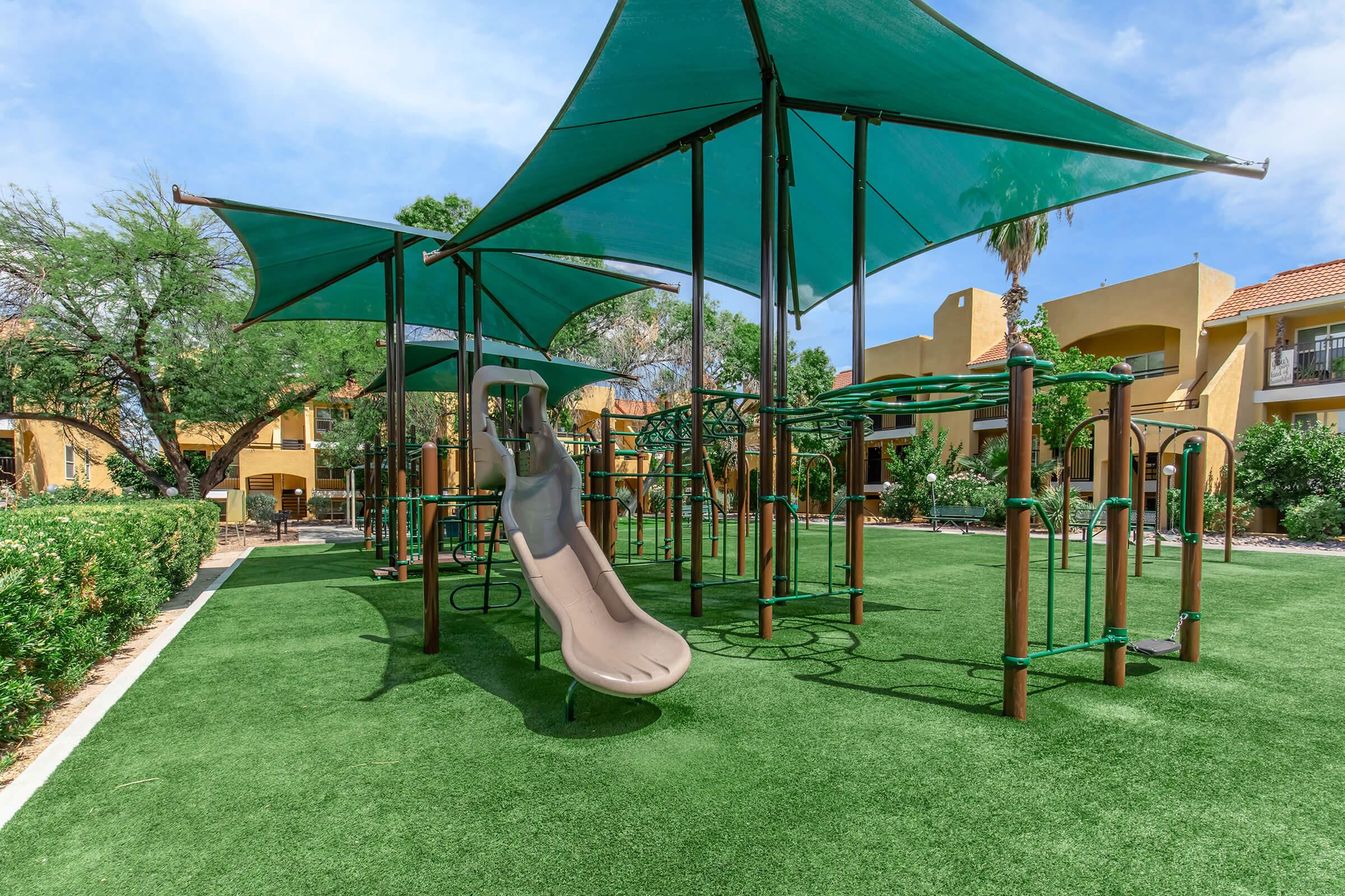 A playground area featuring a curved slide, climbing structures, and green shade canopies. The ground is covered with artificial grass, surrounded by shrubs and trees, set against a backdrop of residential buildings in a sunny environment.