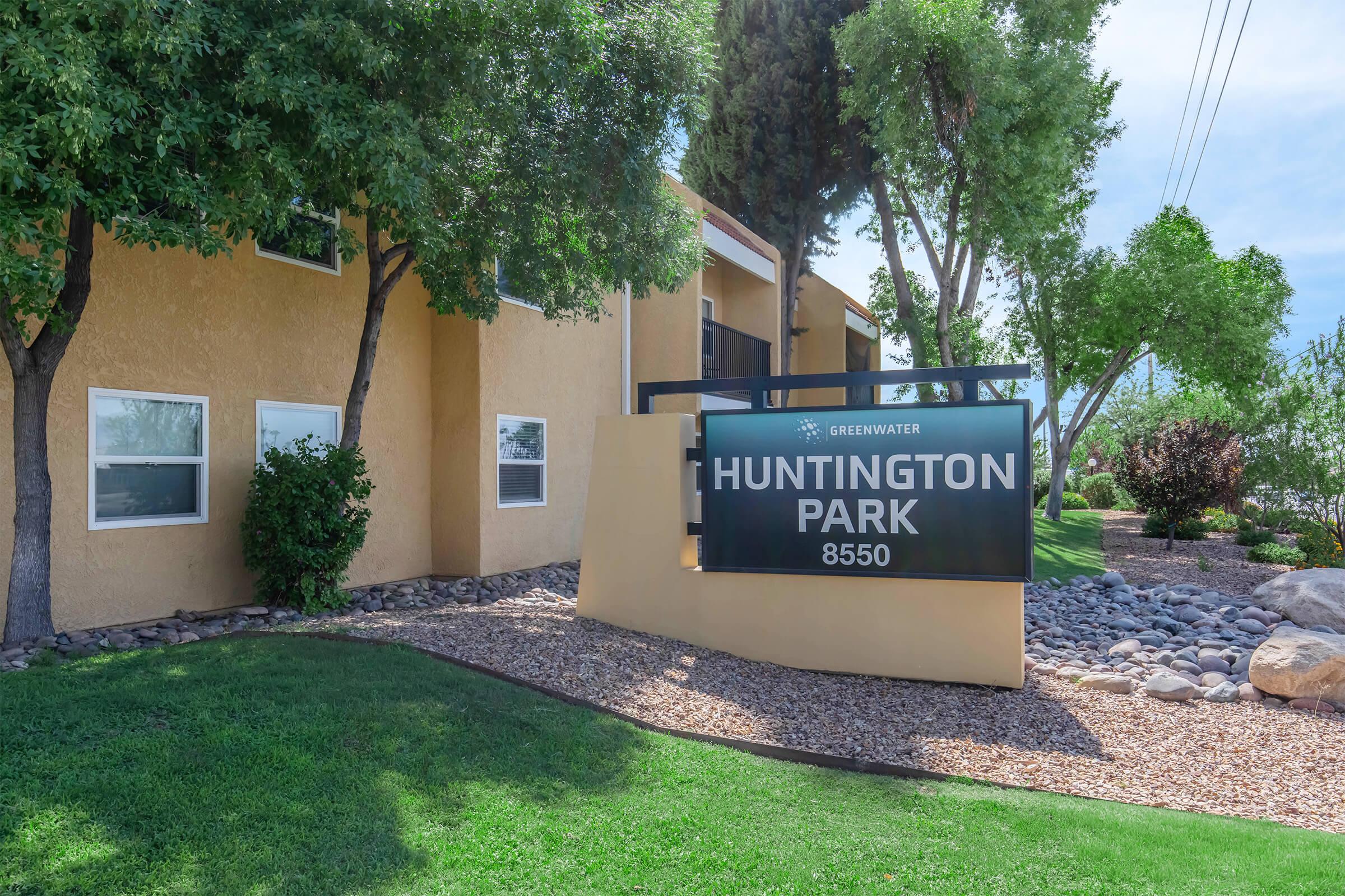 Sign for Huntington Park located at 8550, with a landscape featuring grass, trees, and decorative rocks. The building is painted in a warm color, and the area is well-maintained with foliage.