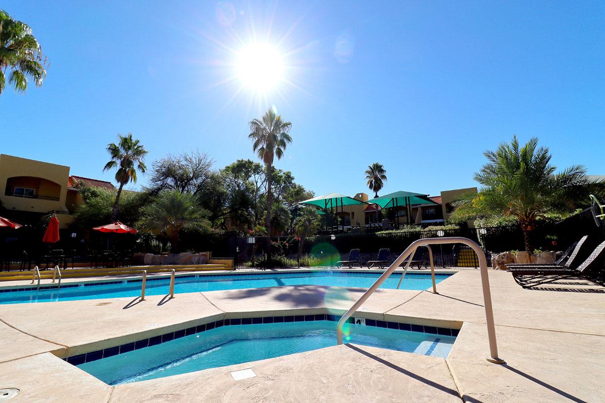 A sunny poolside scene featuring a clear blue sky, a shining sun, and palm trees. The pool has a unique octagonal shape with steps leading in and is surrounded by lounge chairs and umbrellas. Lush greenery adds to the tranquil atmosphere of the setting.