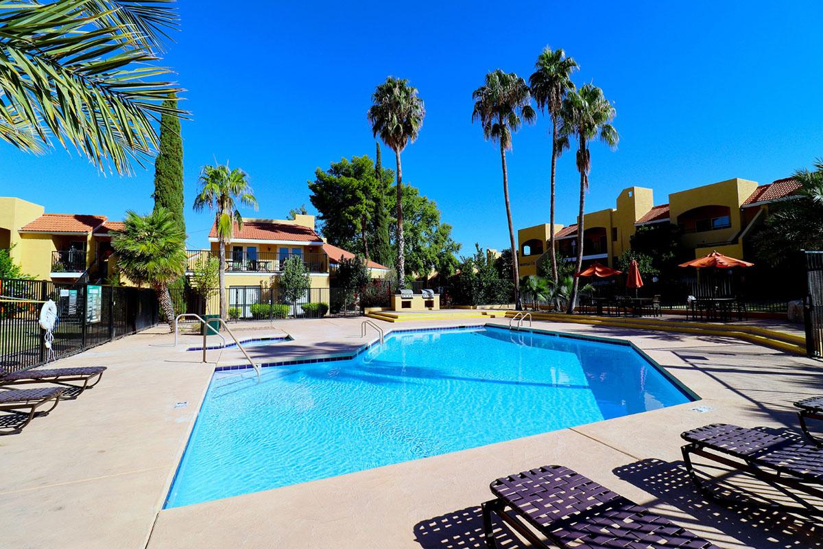 A bright pool area surrounded by palm trees, lounge chairs, and yellow buildings in the background. The clear blue sky enhances the serene atmosphere, making it a perfect spot for relaxation and leisure.
