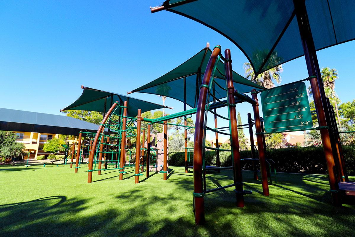 A playground with climbing structures and shade canopies under a clear blue sky. The ground is covered with green grass, and palm trees are visible in the background, creating a vibrant outdoor play area.
