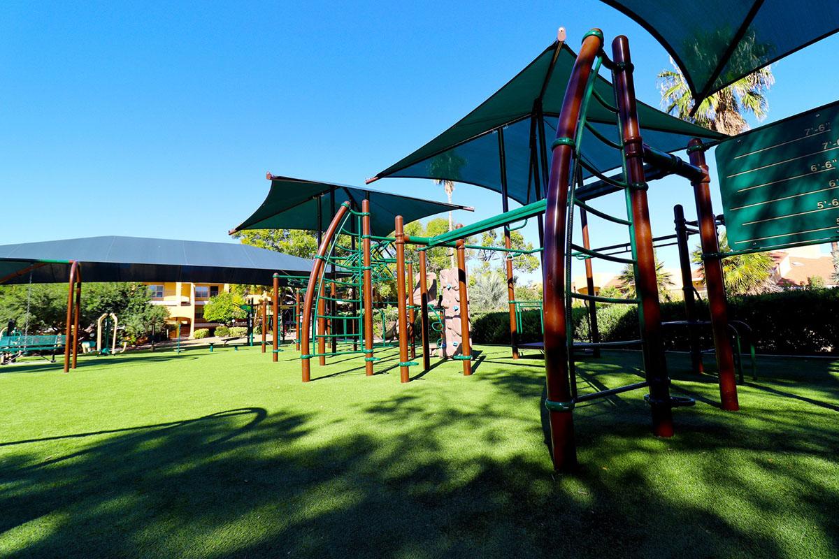 A colorful outdoor playground equipped with climbing structures and shaded areas, set on a grassy surface. Clear blue sky overhead and palm trees in the background, creating a fun and inviting space for children to play and explore.