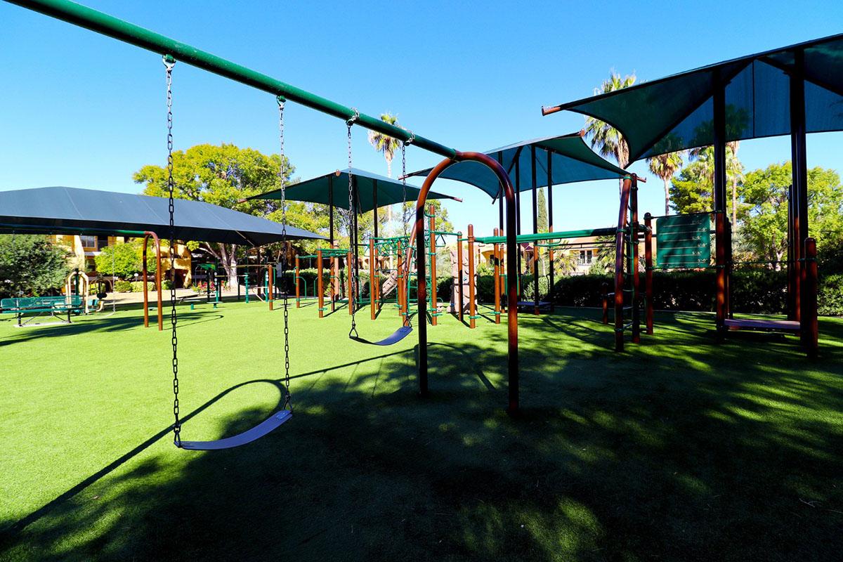 A playground area featuring swings and shaded structures, set on green grass under a clear blue sky. Palm trees are visible in the background, creating a bright and inviting atmosphere for children to play.