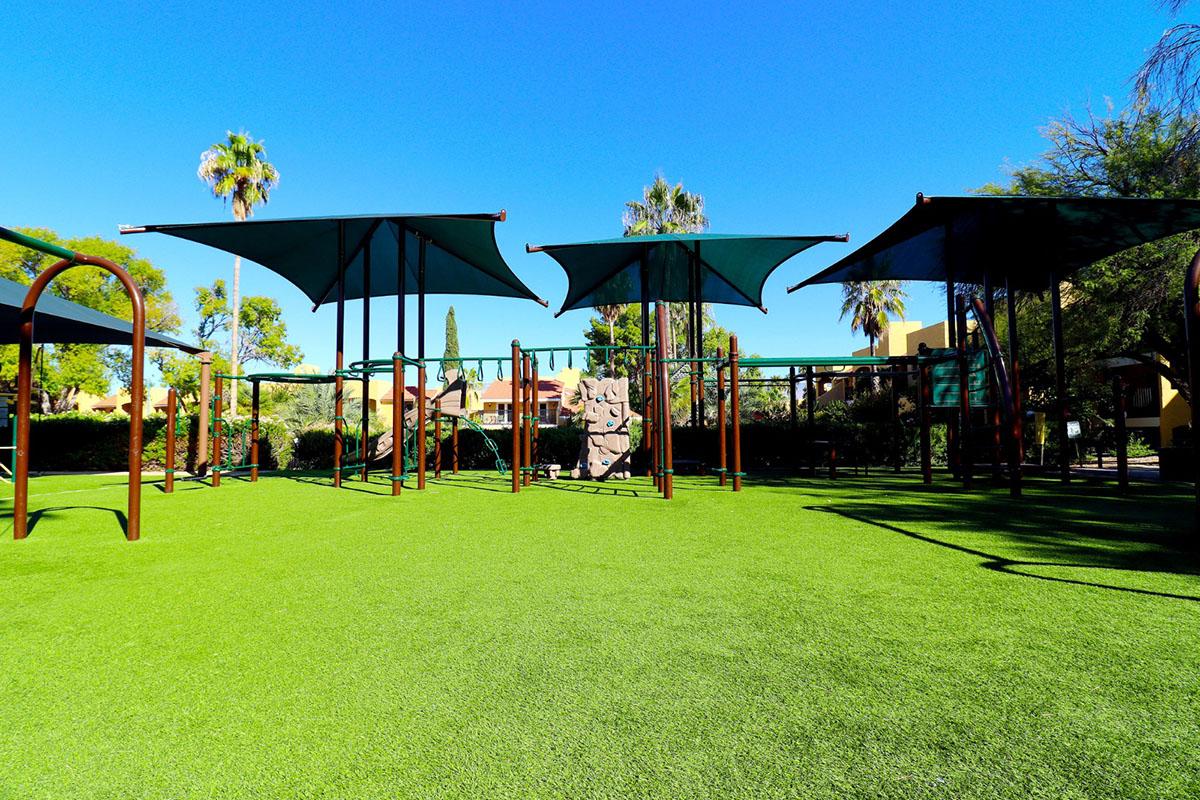 A playground scene featuring green synthetic grass, with several metal structures and shaded areas created by large, angular canopies. The background includes palm trees and a sunny blue sky, providing a bright and inviting setting for outdoor play.