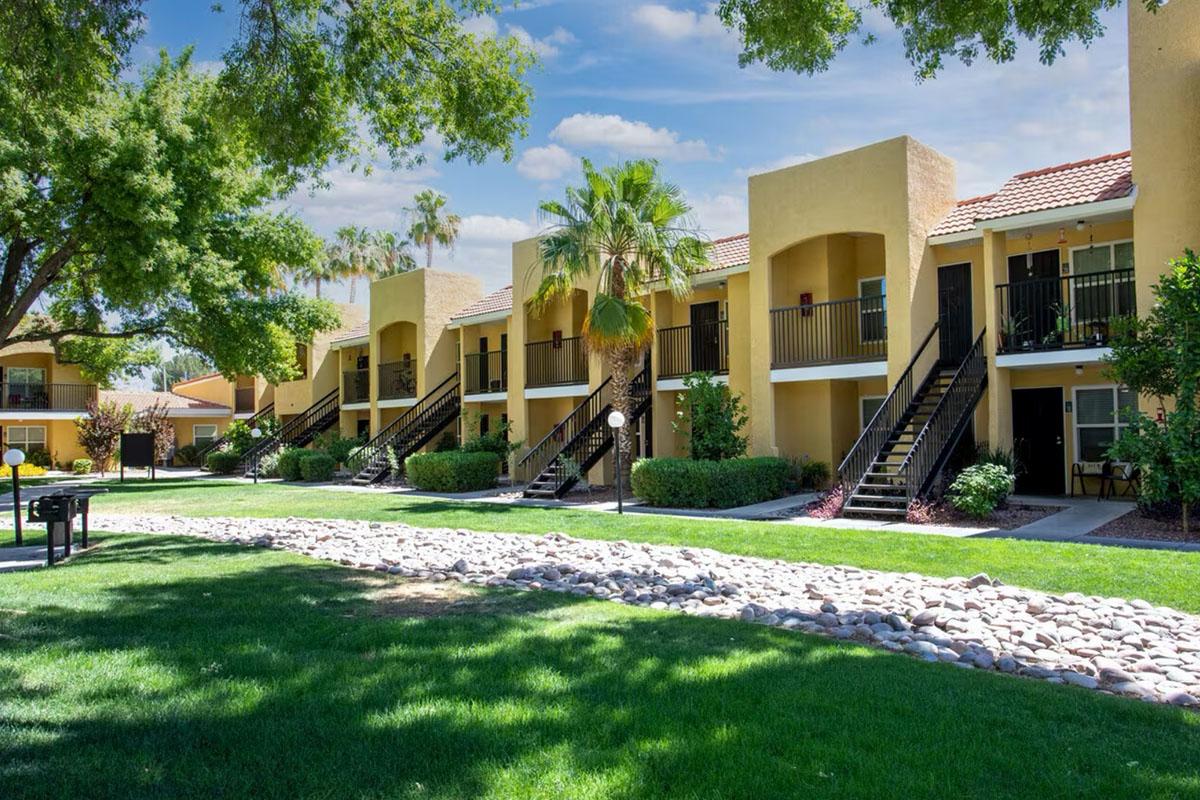 Two-story apartment buildings with balconies, surrounded by lush green grass and palm trees. A pebble pathway runs through the area, leading past the buildings. The sky is bright with a few clouds, creating a sunny atmosphere in a residential community setting.