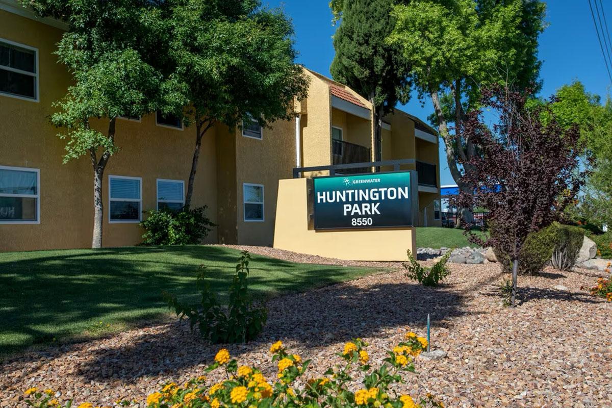 Sign for Huntington Park located at 8550, in front of a yellow apartment building. The area features green grass and decorative shrubs, with colorful flowers in the foreground. Bright blue sky enhances the inviting atmosphere of the property.