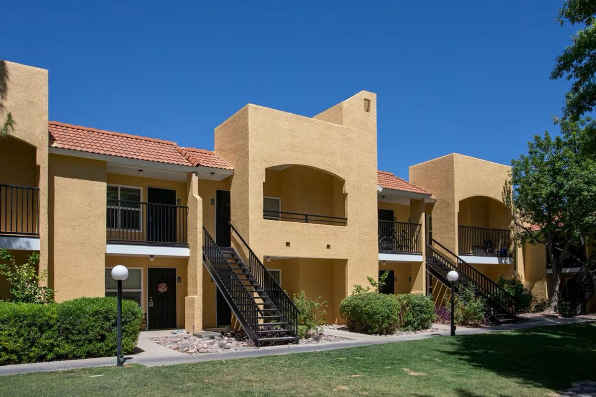 Two-story apartment buildings with a tan exterior and red tile roofs, featuring black metal railings on staircases. The buildings are surrounded by green grass and shrubs, with a clear blue sky in the background. Two decorative light posts are visible in the foreground.
