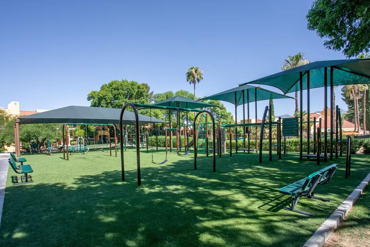 A spacious playground featuring green synthetic grass, shaded areas with large canopies, swings, climbing structures, and benches. Surrounding palm trees and a clear blue sky enhance the inviting atmosphere for children to play and socialize.