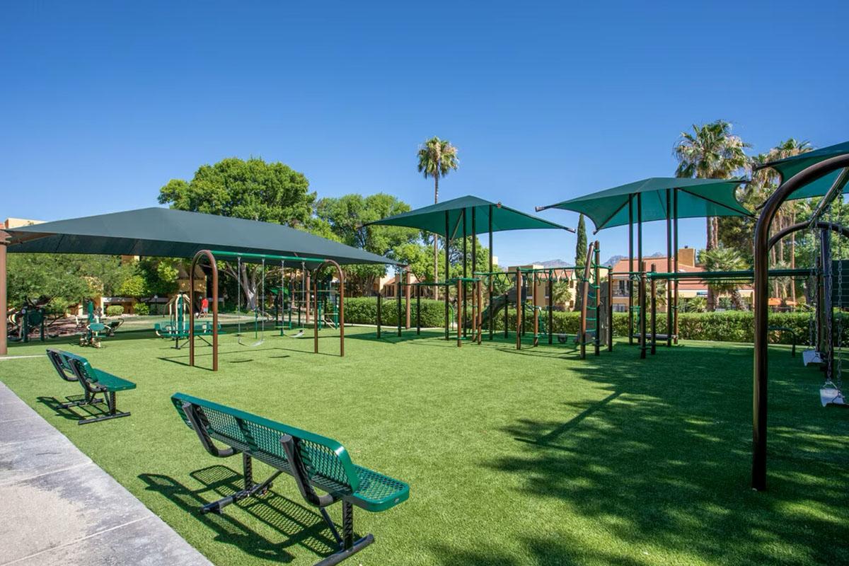 A sunny playground with green artificial turf, featuring climbing structures and swings under large green shade canopies. Benches are positioned along the path, surrounded by palm trees and lush greenery, creating a welcoming outdoor recreational space.