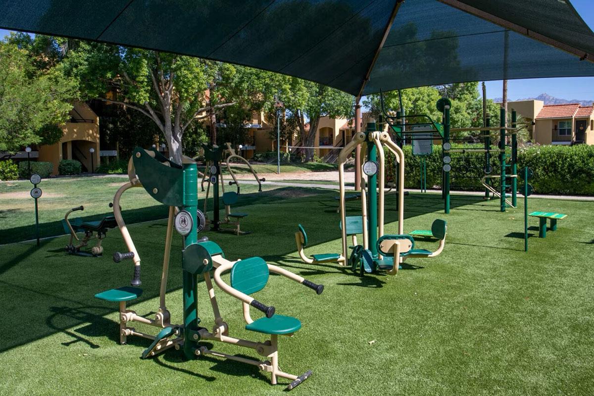 Outdoor fitness area featuring various exercise machines under a shaded canopy. The space is surrounded by green grass and trees, with nearby buildings in the background. Ideal for outdoor workouts in a community setting.