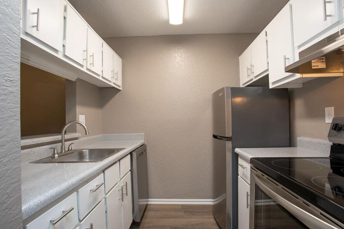 A modern kitchen featuring white cabinets, a stainless steel refrigerator, a silver dishwasher, and a sleek countertop. The space includes a sink with a faucet, an electric stovetop, and light gray walls, illuminated by a ceiling light. The flooring is laminate wood, creating a warm ambiance.