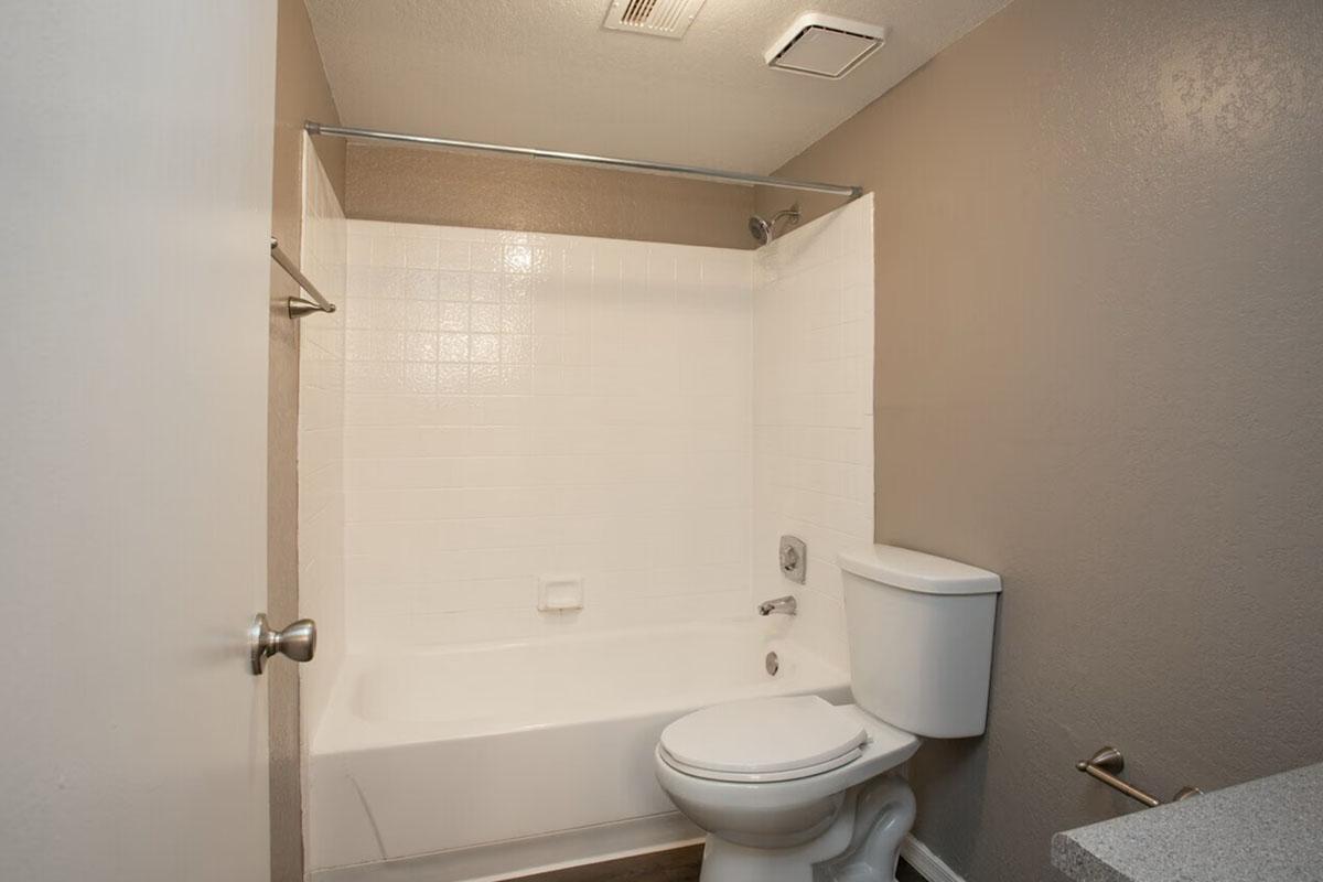 A clean, modern bathroom featuring a white bathtub with a shower, a toilet, and a simple countertop. The walls are a light brown color, and there is a small ceiling light above. The overall design is minimalistic and functional.