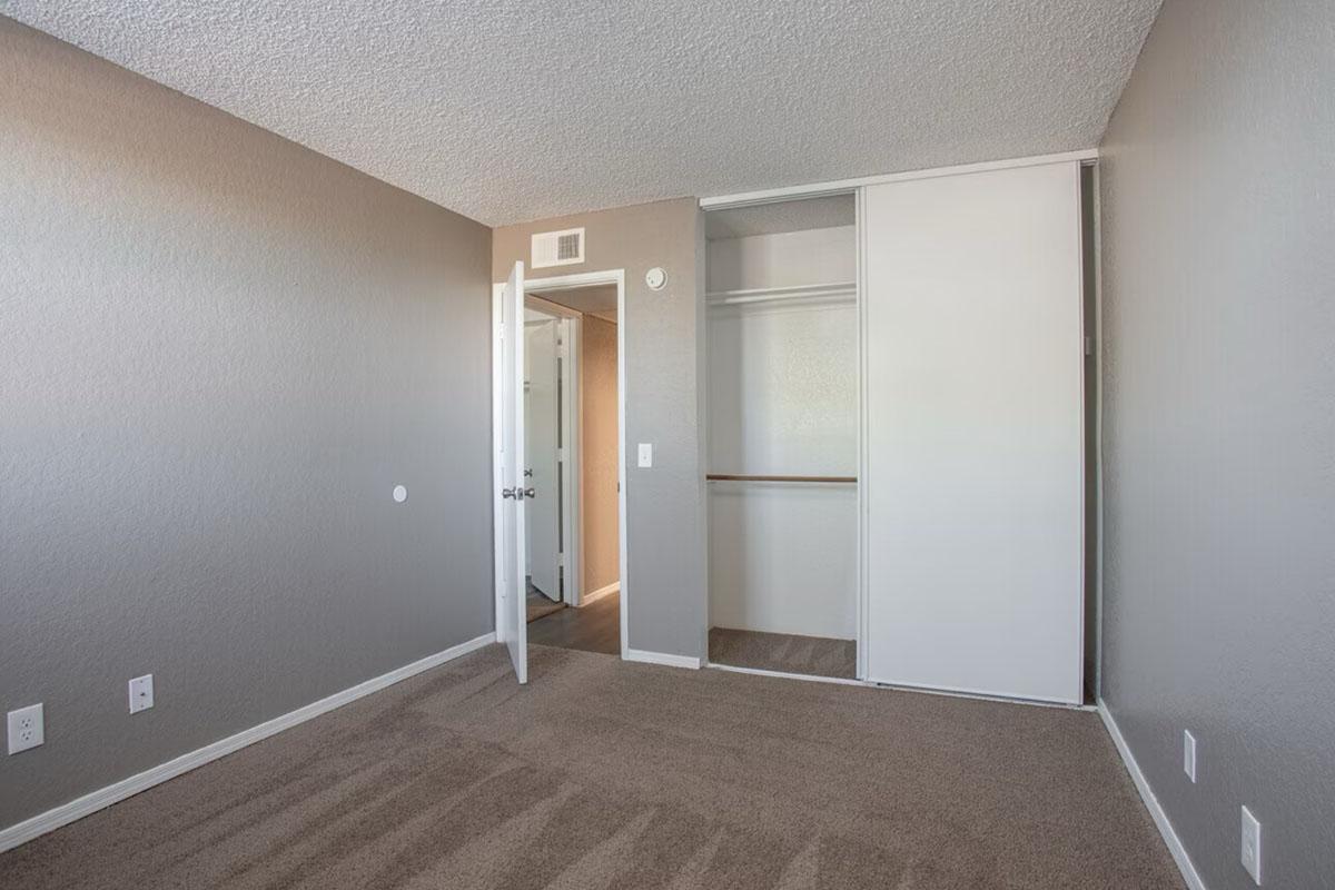 A vacant room featuring light gray walls and a carpeted floor. There is a closet with sliding doors on one side, and a white door leading to another room is slightly ajar. The overall atmosphere is simple and unadorned, ideal for potential customization.