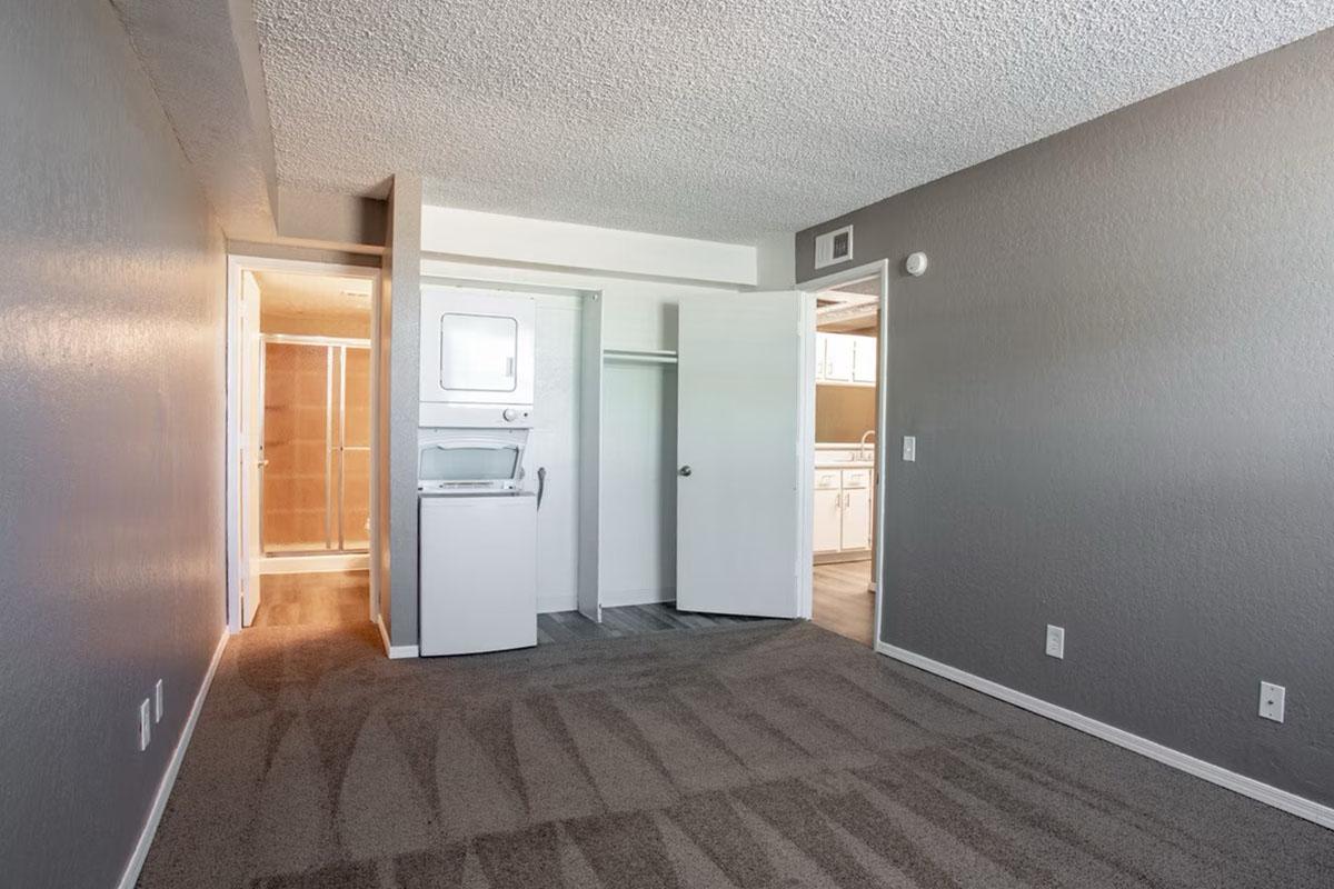 A spacious room with gray walls and carpet. A doorway leads to a bathroom area, while another door opens to a closet with a stacked washer and dryer. The lighting is soft, creating a cozy atmosphere. The room appears clean and well-maintained, suitable for various uses.
