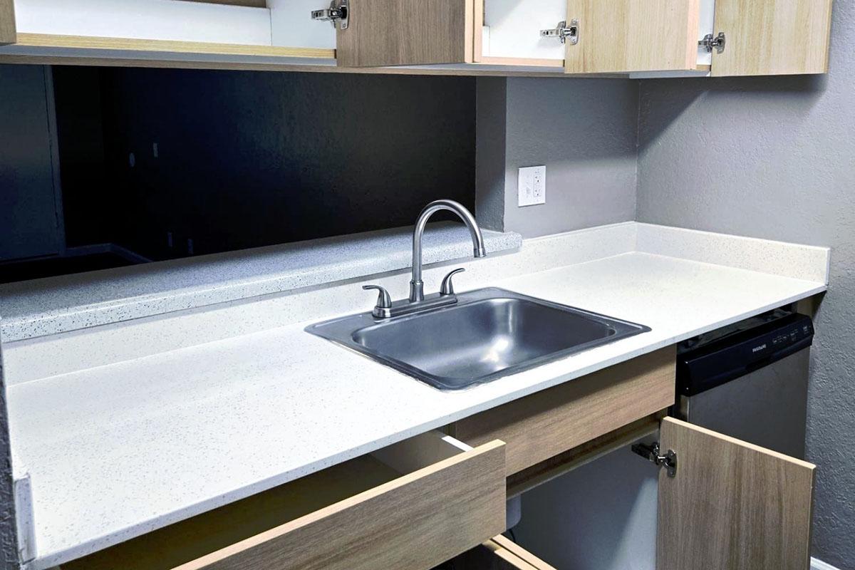 A modern kitchen countertop featuring a stainless steel sink, with an open drawer below and another drawer beside it. The cabinetry is light wood, and the countertop is white, creating a clean and contemporary look.