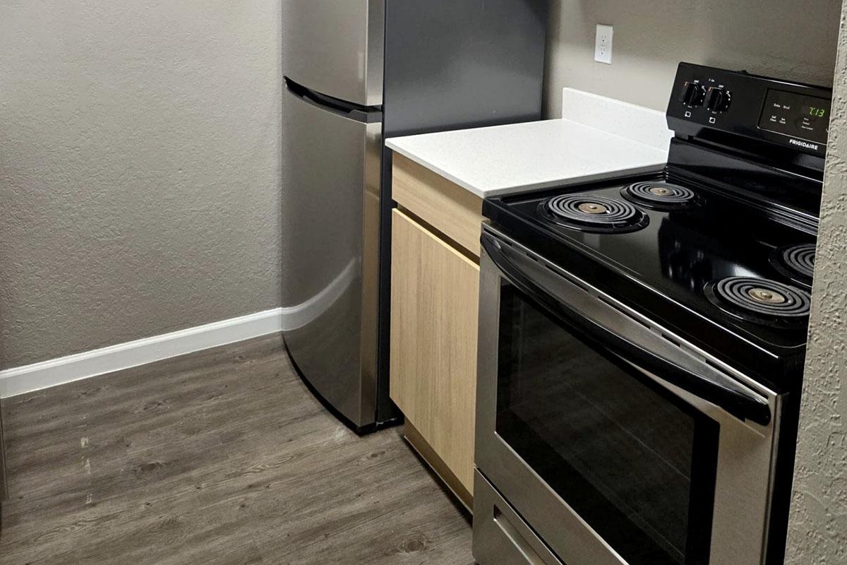 A modern kitchen featuring a stainless steel refrigerator, a black electric stove, and a light wood cabinet. The countertop is white with a smooth finish, and the floor has a wood-like design. The walls are painted in a neutral color, enhancing the contemporary look of the space.