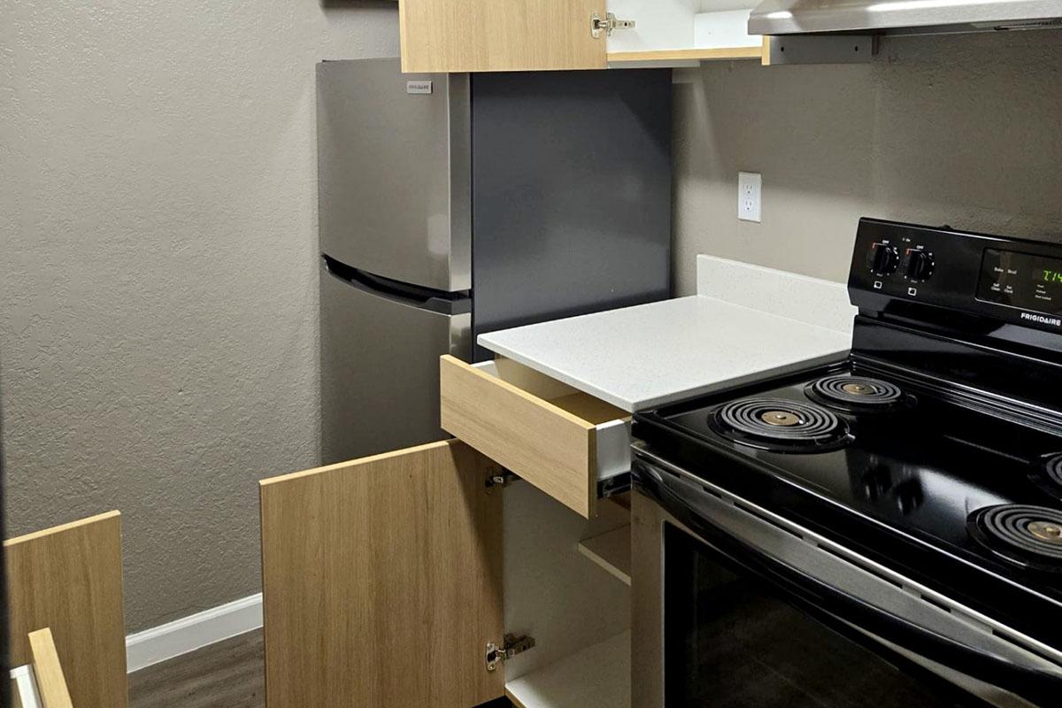 A modern kitchen featuring a black stove with four burners, a silver refrigerator, and light wood cabinetry. The countertop is white, and some drawers are open, displaying a tidy interior. The walls are painted in a neutral color, creating a clean and functional space.