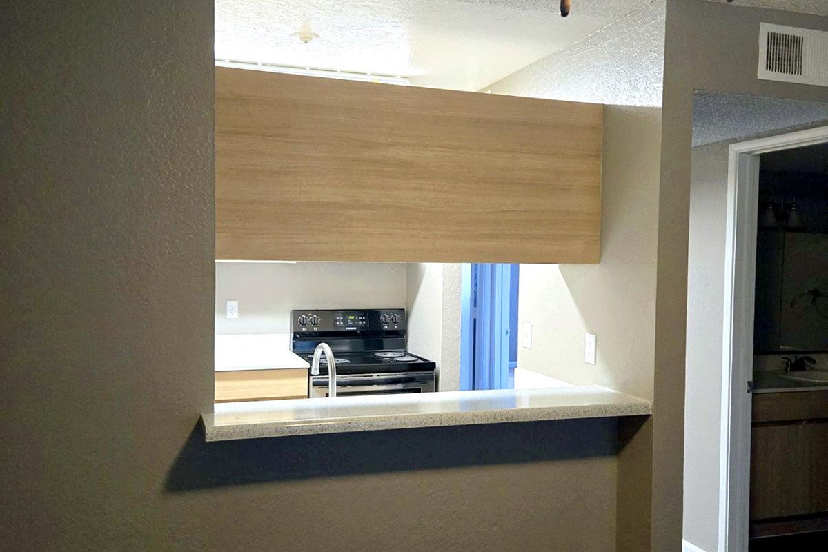 View of a modern kitchen with a beige and wood design. An open counter connects the kitchen to the living area. The kitchen features a black stove, white cabinets, and light-colored countertops. There is a blue accent wall seen through the opening, enhancing the spacious feel of the layout.