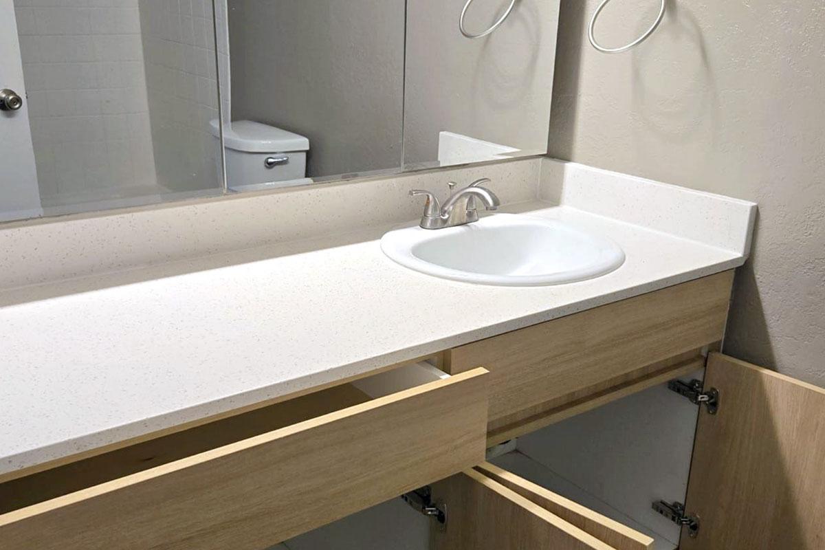 A clean bathroom sink area featuring a white countertop, a single sink with a faucet, and a large mirror above. The wooden cabinetry below has open drawers revealing empty storage space. In the background, a white toilet is partially visible. The overall color scheme is neutral and modern.