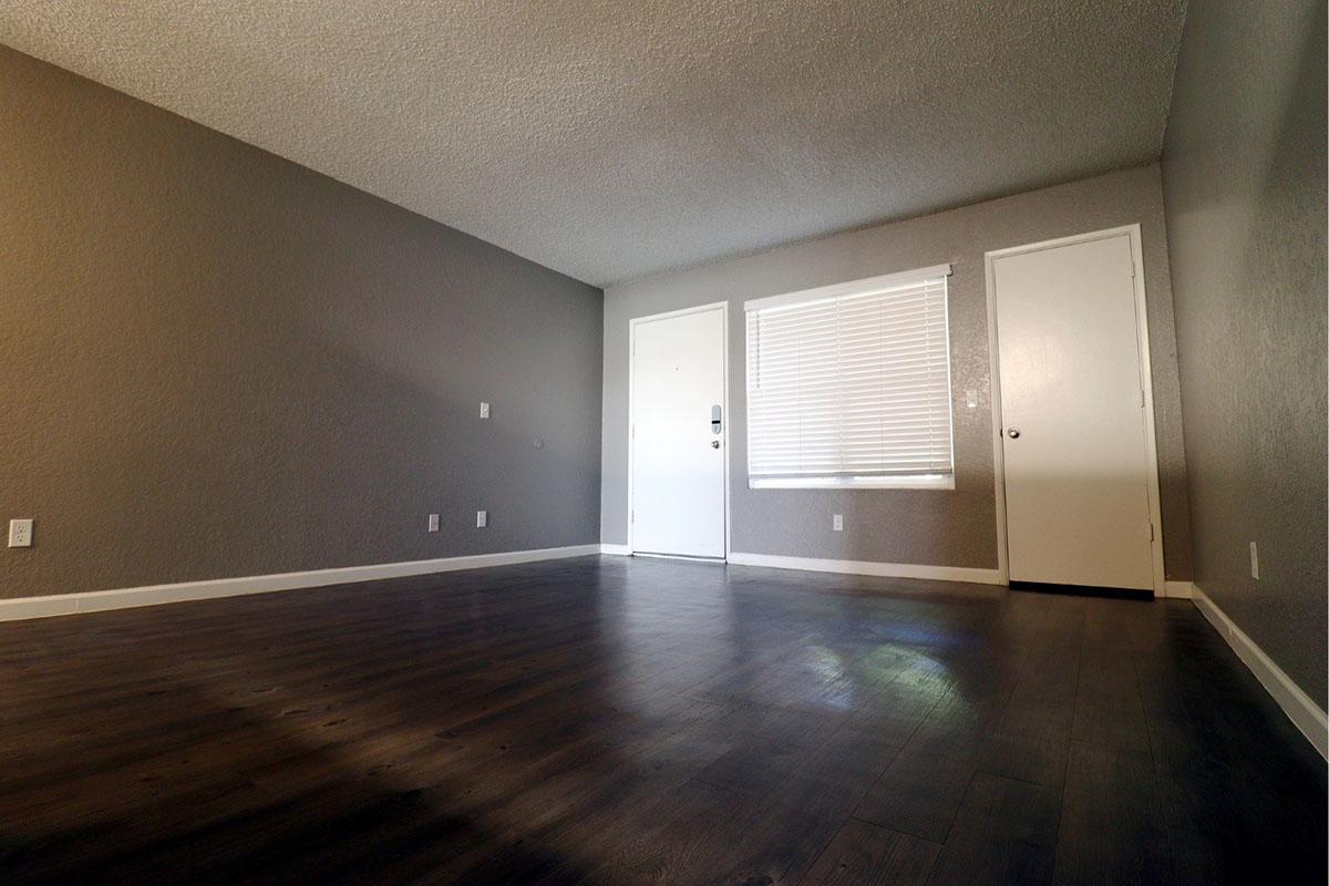 A spacious, empty room with light gray walls and dark wood flooring. There is a door on the left, a window with blinds allowing natural light on the right, and a door opposite the entrance. The room appears well-lit and features a textured ceiling.