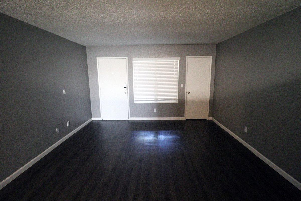 An empty, well-lit room with gray walls and dark laminate flooring. There are two closed doors on the right side and a window with white blinds on the left side. The space appears clean and minimalistic, suitable for various uses or decoration.