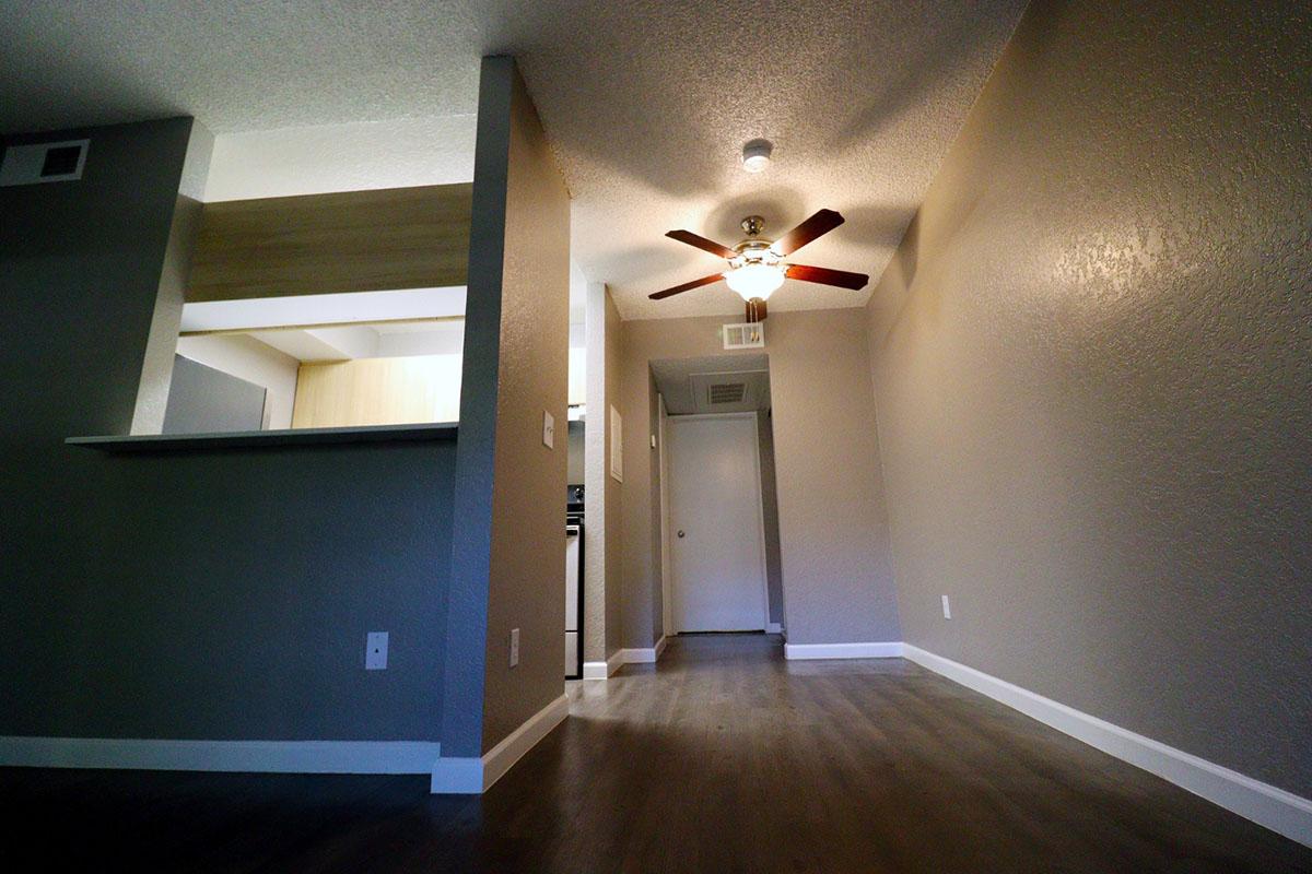 Interior view of a hallway in an apartment, featuring a ceiling fan with wooden blades, light-colored walls, and laminate flooring. The open space leads to a kitchen area on the left, with a doorway visible at the end of the hall. The overall ambiance is bright and modern.