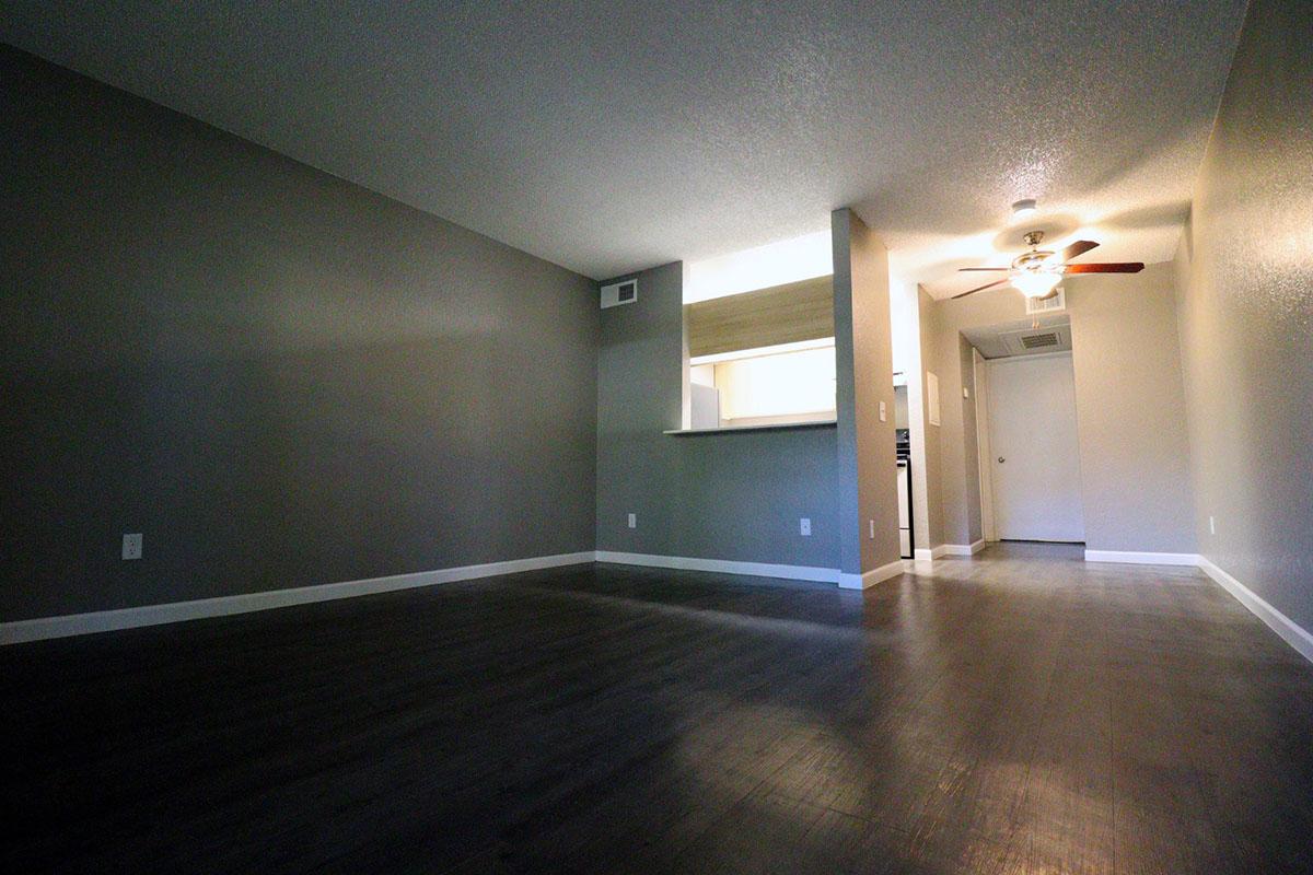 A spacious, modern living room with dark wood flooring and gray walls. A small kitchen area is visible in the background with light-colored cabinetry. A ceiling fan is mounted above, and a door to another room can be seen in the distance, creating an open and inviting atmosphere.
