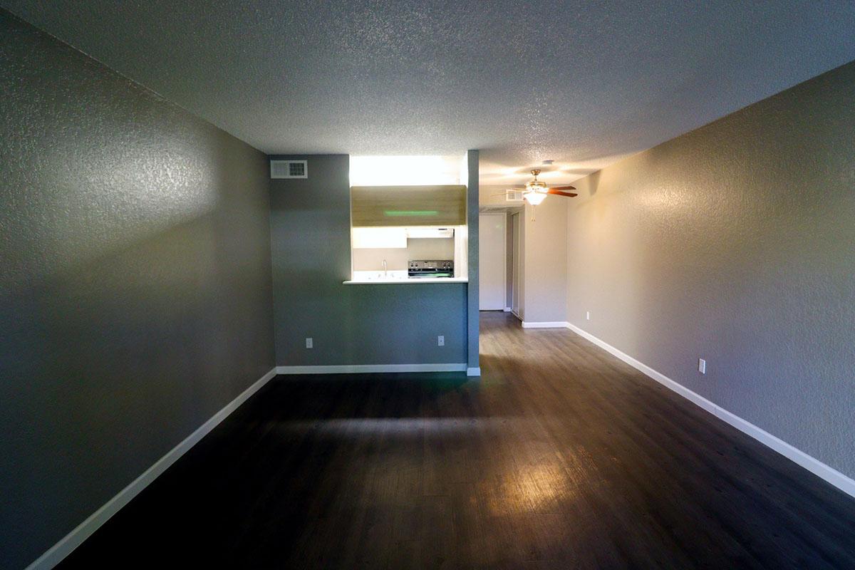 A spacious, empty living area with gray walls and dark laminate flooring. To the left, an open kitchen area features a counter with a bar height, illuminated by light from the kitchen. A ceiling fan is visible, and a doorway leads to another room, creating a sense of openness.