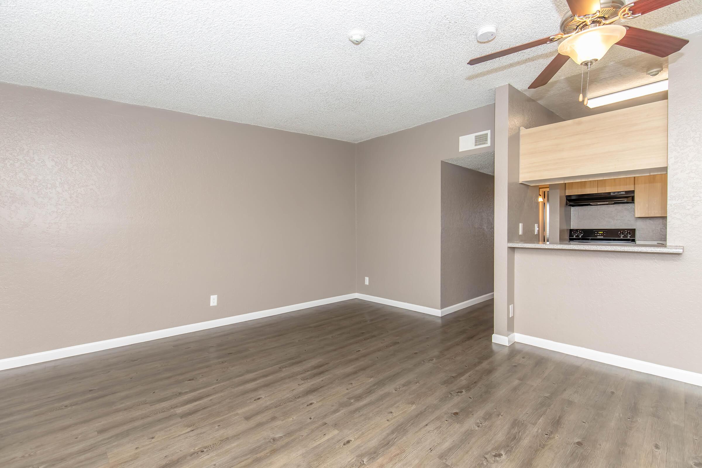 Spacious, empty living area with light wood flooring and neutral walls. A ceiling fan hangs from the textured ceiling. To the right, a small kitchen area with a bar counter is visible, featuring light wood cabinetry and a black stove. The overall atmosphere is bright and inviting.