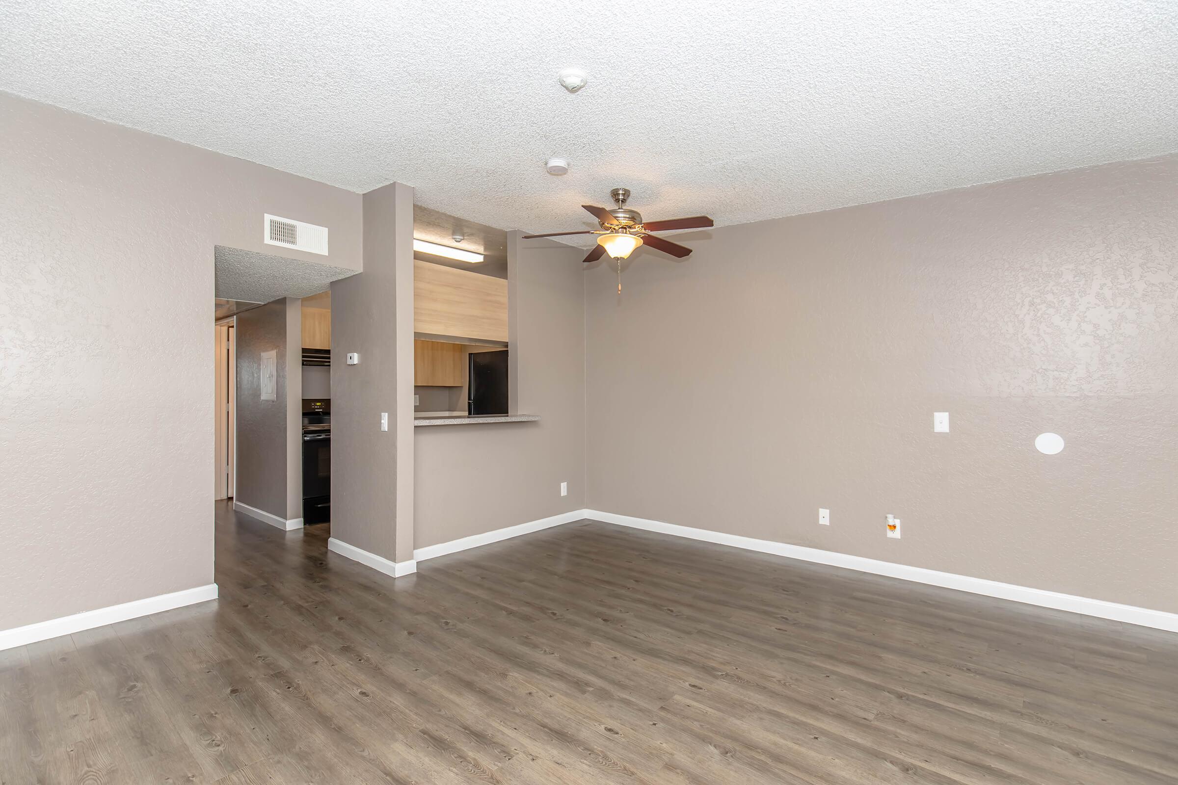 A spacious room featuring light-colored walls and a ceiling fan. There is a bar-height counter leading to a kitchen area with dark appliances visible. The floor is a light wood laminate, and the room has ample natural light from a nearby window.