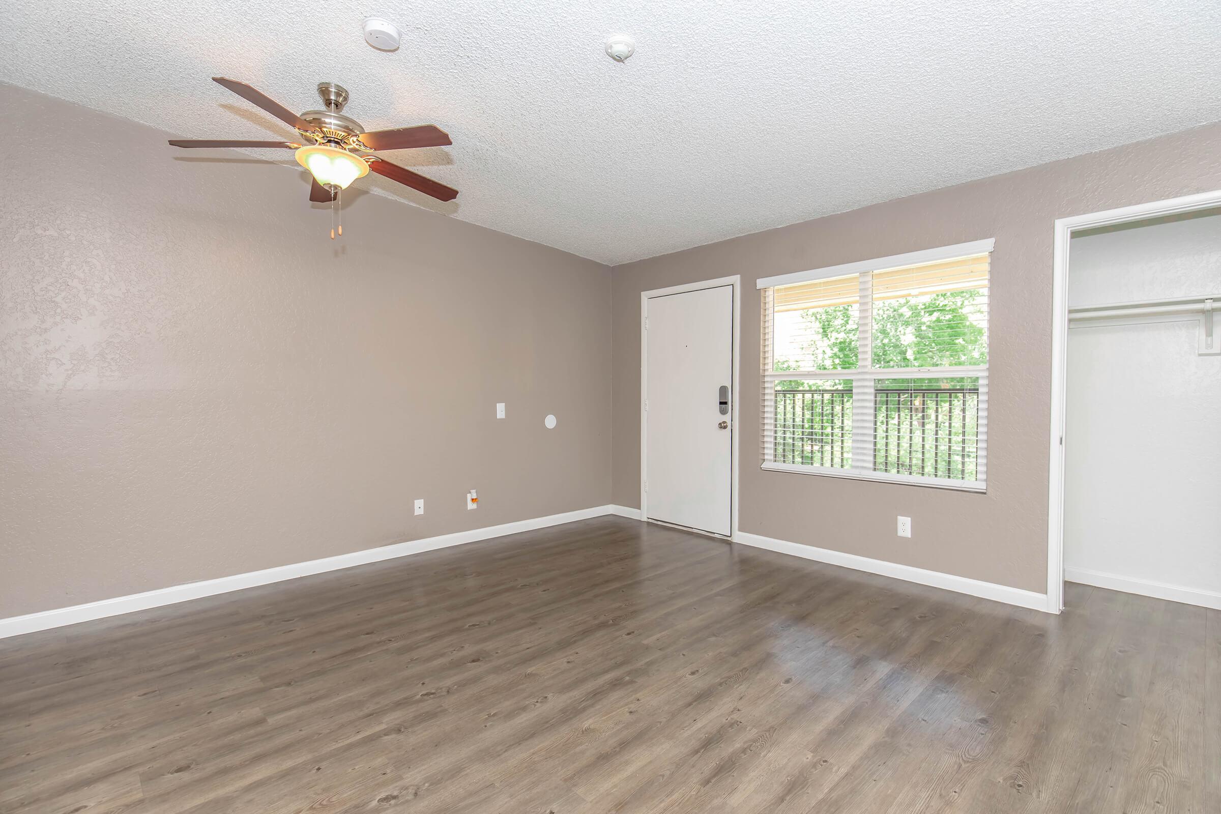 A spacious room with a light brown textured wall, wooden-style flooring, and a ceiling fan. There is a door leading outside, a window allowing natural light, and a closet space on one side. The room appears bright and inviting, perfect for various uses.